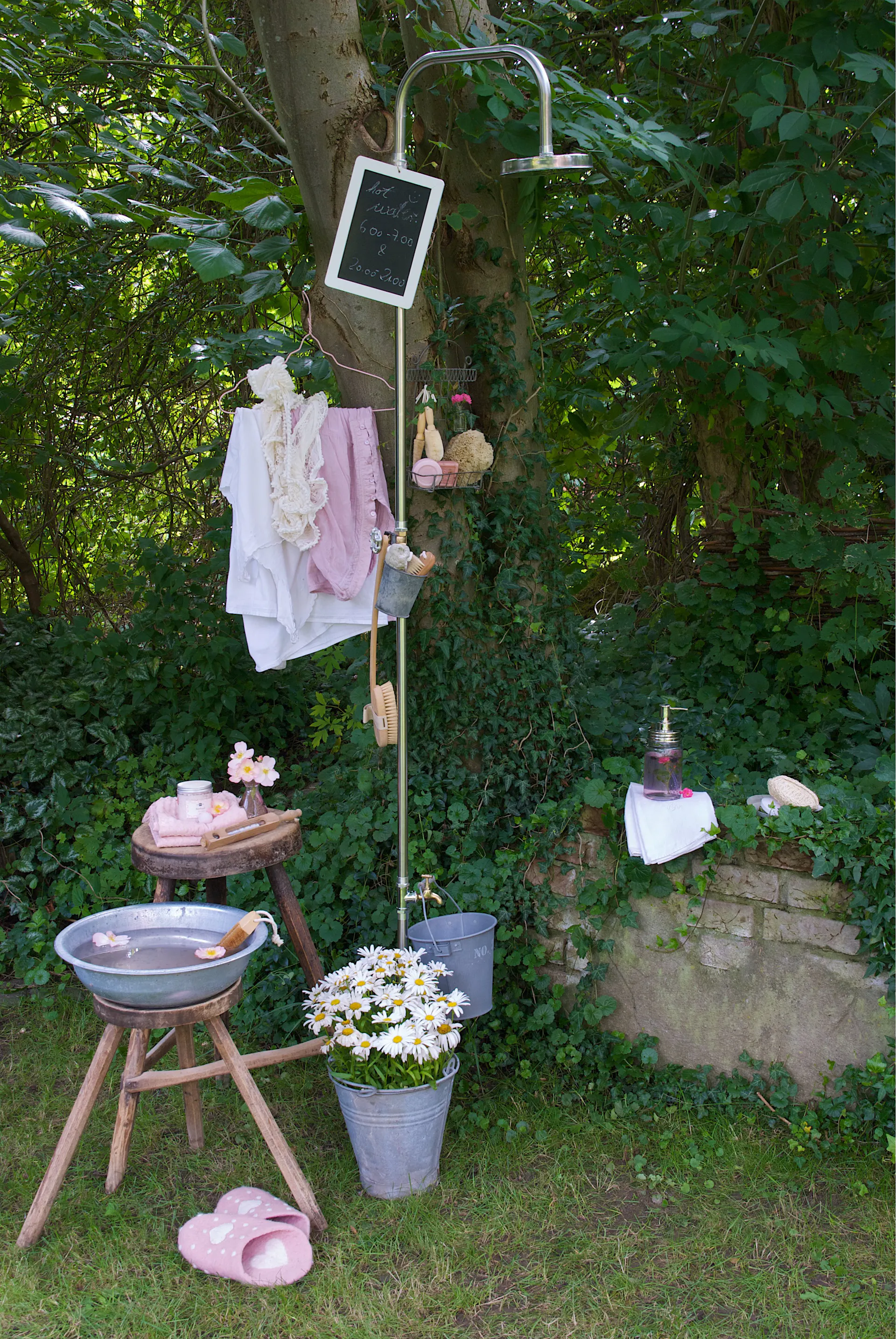 Doccia da giardino mobile fissata a un tronco d’albero nel giardino con accanto accessori da doccia e vestiti