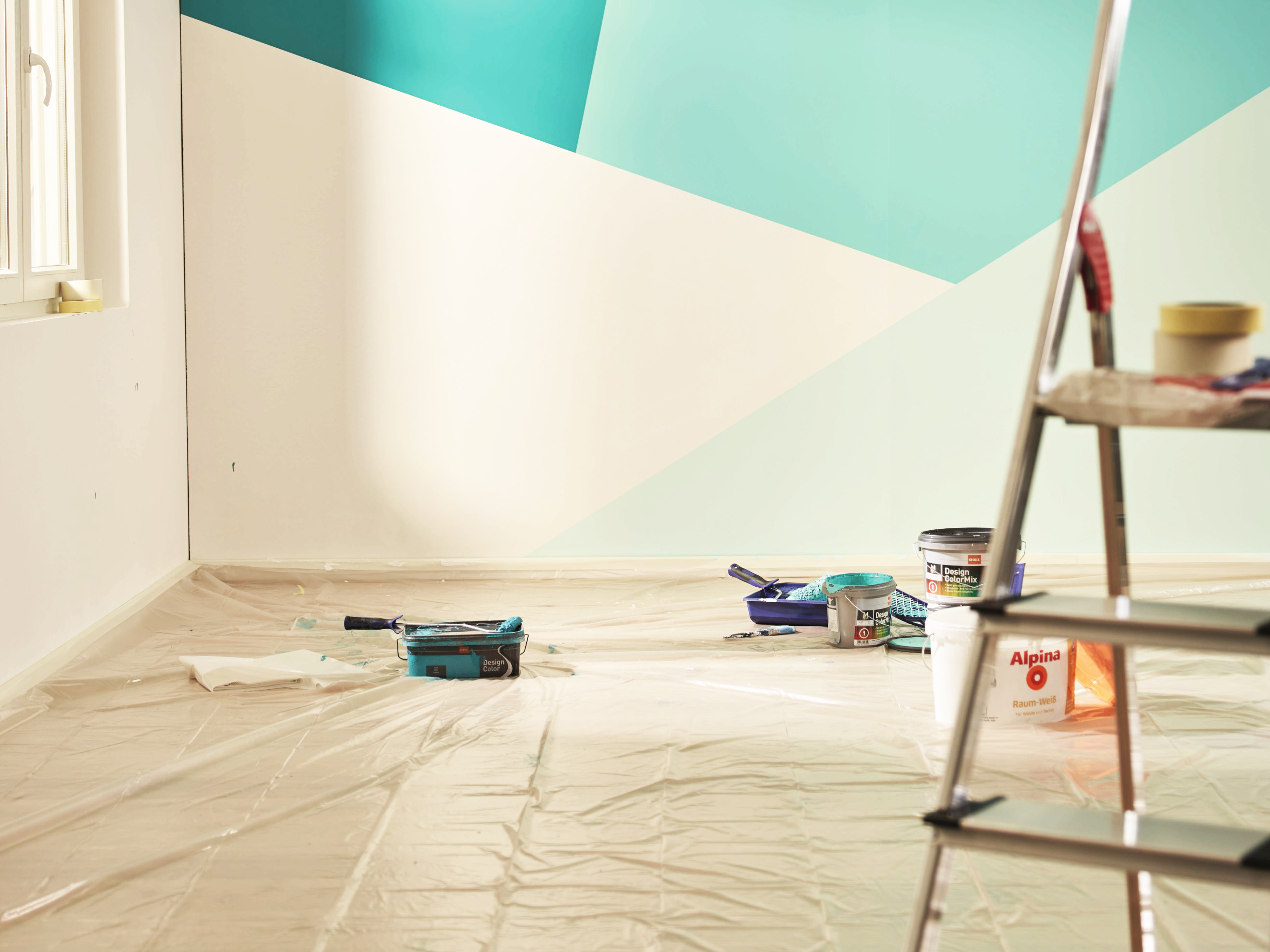 Mur peint en différents tons bleus et motifs géométriques, film protecteur et seau de peinture sur le sol et escabeau au premier plan