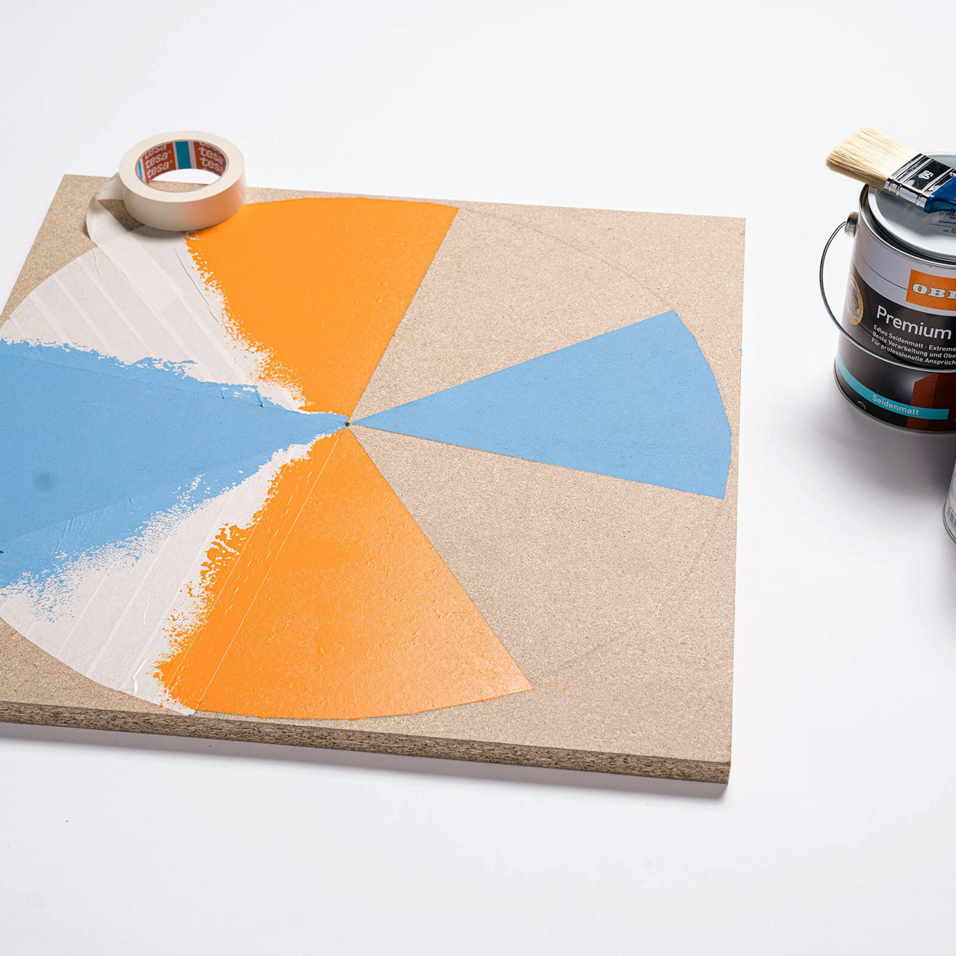 Dreifarbig bemalter Kreis auf einer Spanplatte neben zwei Farbdosen