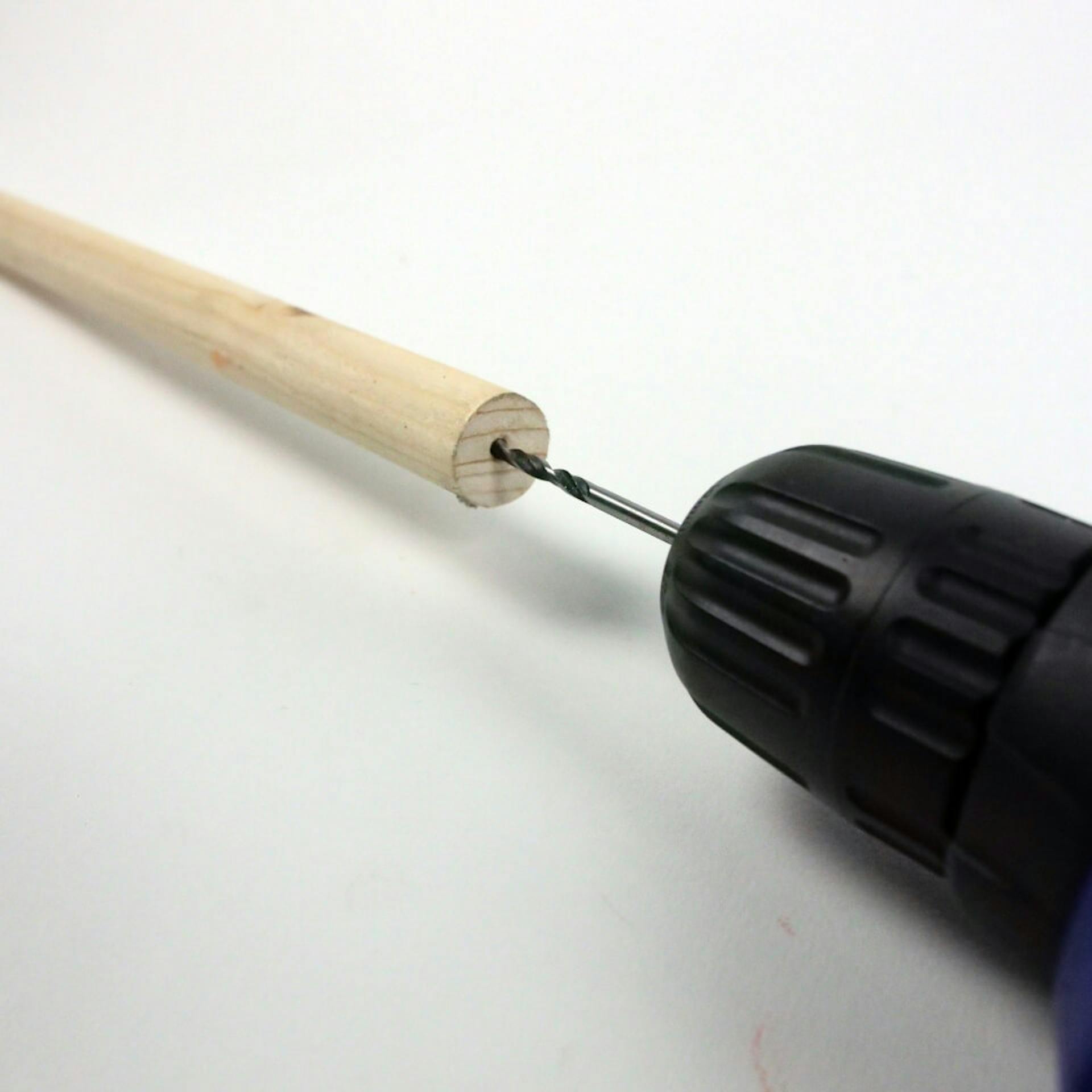 Es ist ein Rundstab aus Holz zu sehen. Daneben ist ein Bohrer zu sehen, welcher in eine Seite des Stabes ein Loch bohrt.
