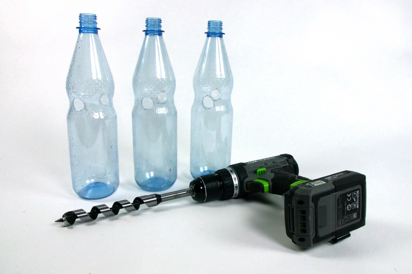 Es sind drei Plastikflaschen mit jeweils zwei Bohrlöchern zu sehen. Vor den Flaschen liegt ein Bohrer.