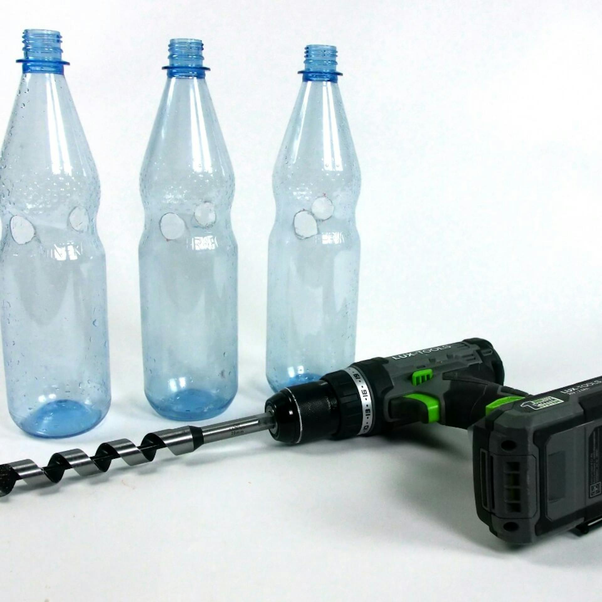 Es sind drei Plastikflaschen mit jeweils zwei Bohrlöchern zu sehen. Vor den Flaschen liegt ein Bohrer.