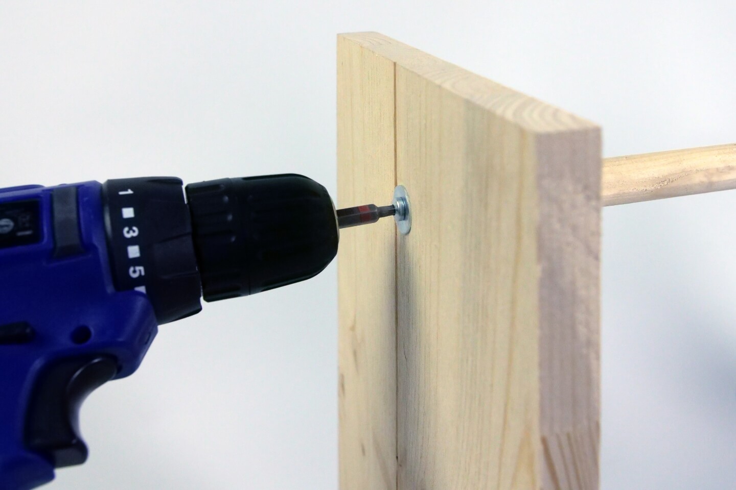 Es ist ein Akkuschrauber zu sehen, welcher ein Holzbrett und einen Rundstab aus Holz zusammenschraubt.