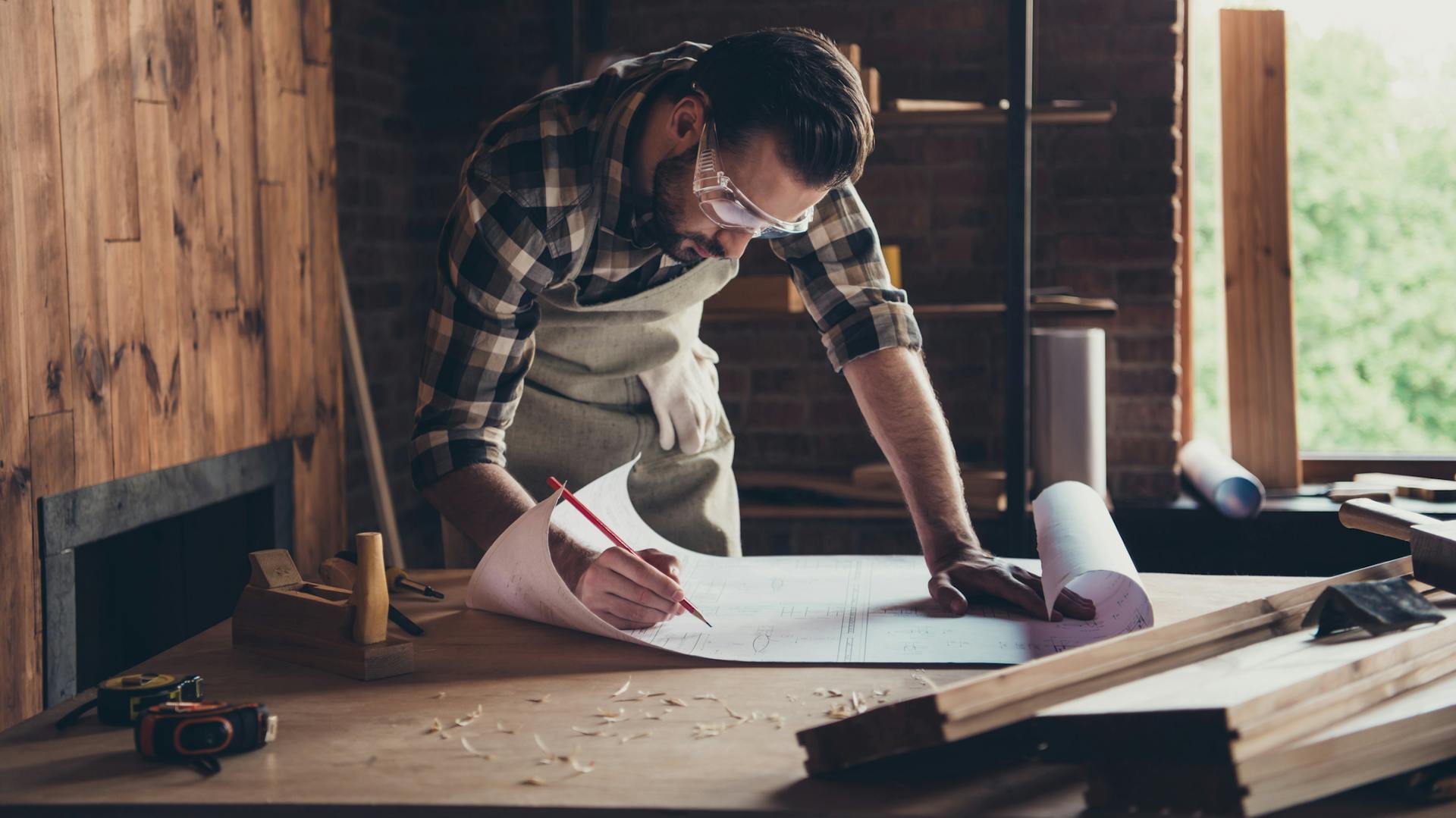 Ein Mann beugt sich über einen Bauplan, in der Hand hält er einen Bleistift, auf dem Tisch liegen Holzspäne.