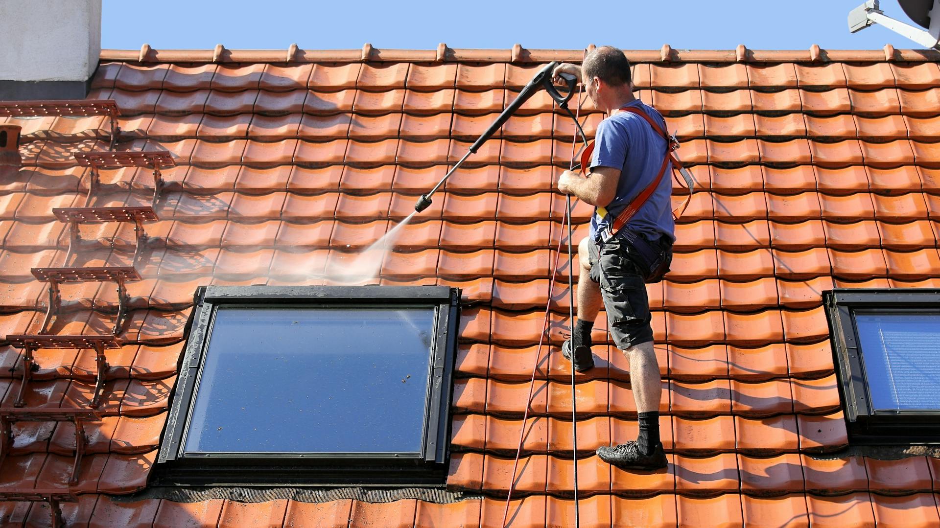 Egy kibiztosított férfi tetőcserepeket tisztít magasnyomású mosóval.