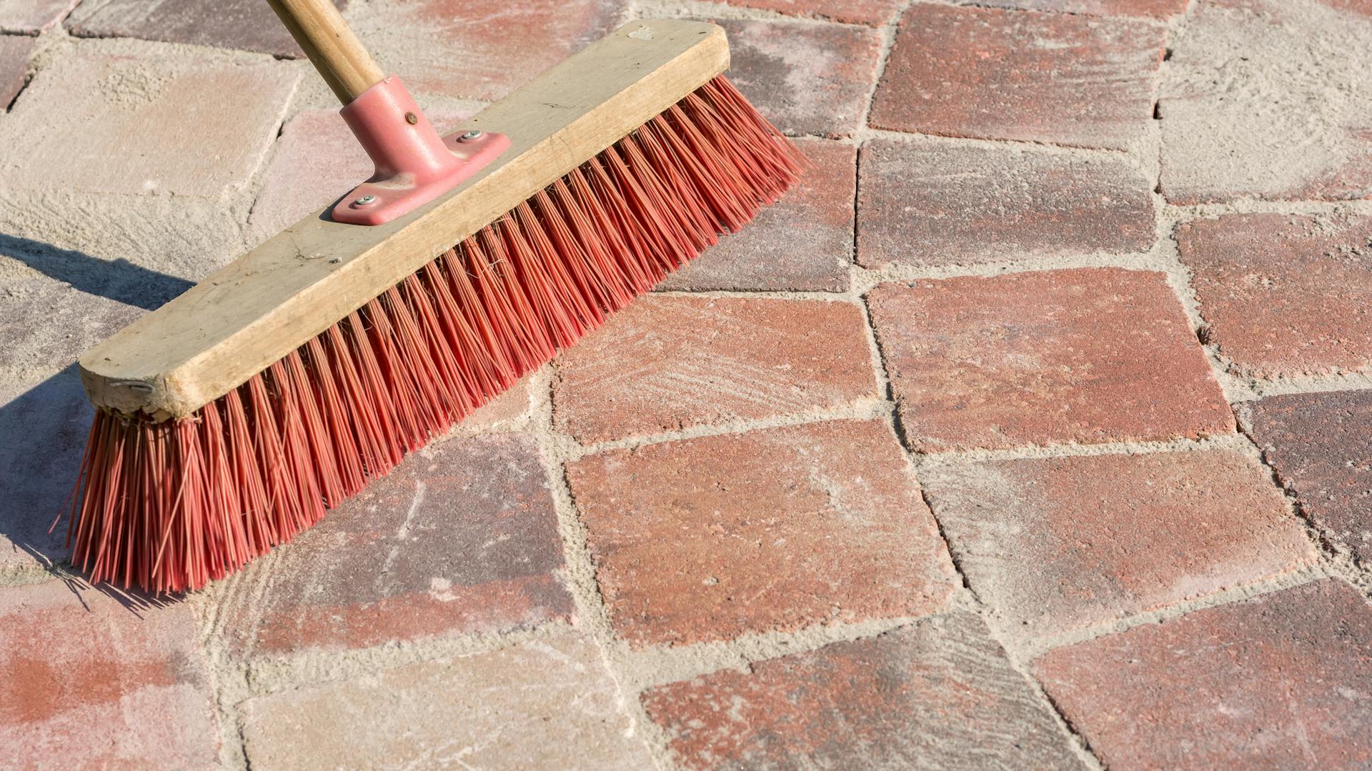 Mit einem Holzbesen mit festen Borsten wird grober Schmutz von Pflastersteinen gefegt.