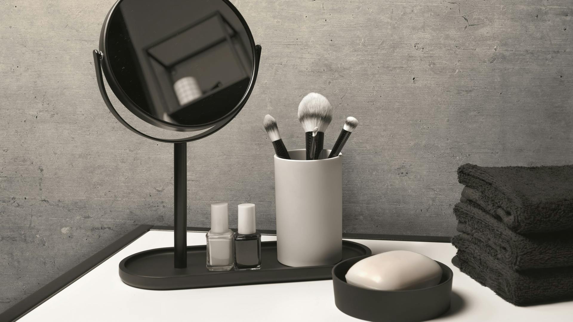 Dunkle und hellgraue Badaccessoires, unter anderem Spiegel, Becher für Schminkpinsel, Seifenschale und Handtücher, für Bad in Betonoptik