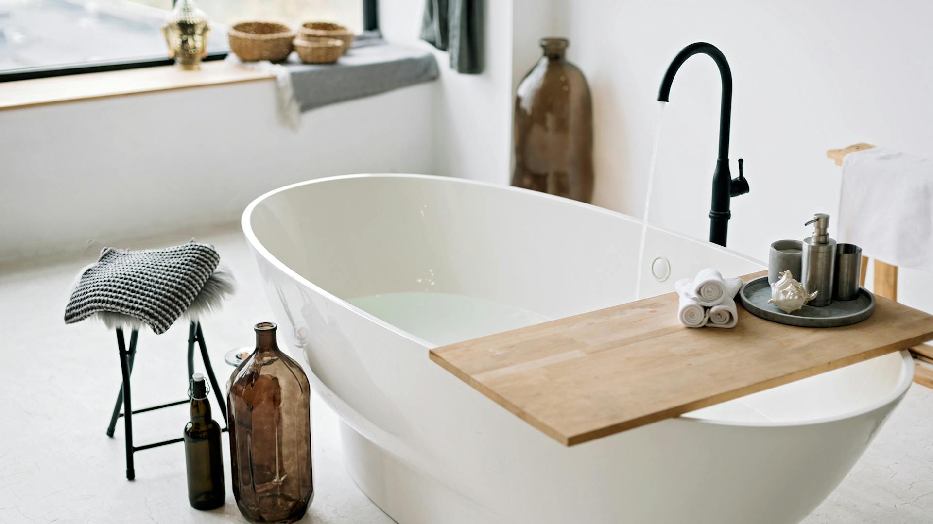 Weiße freistehende ovale Badewanne. Darauf liegt ein Brett mit verschiedenen Badutensilien. Neben der Wanne steht ein Hocker und leere Vasen.