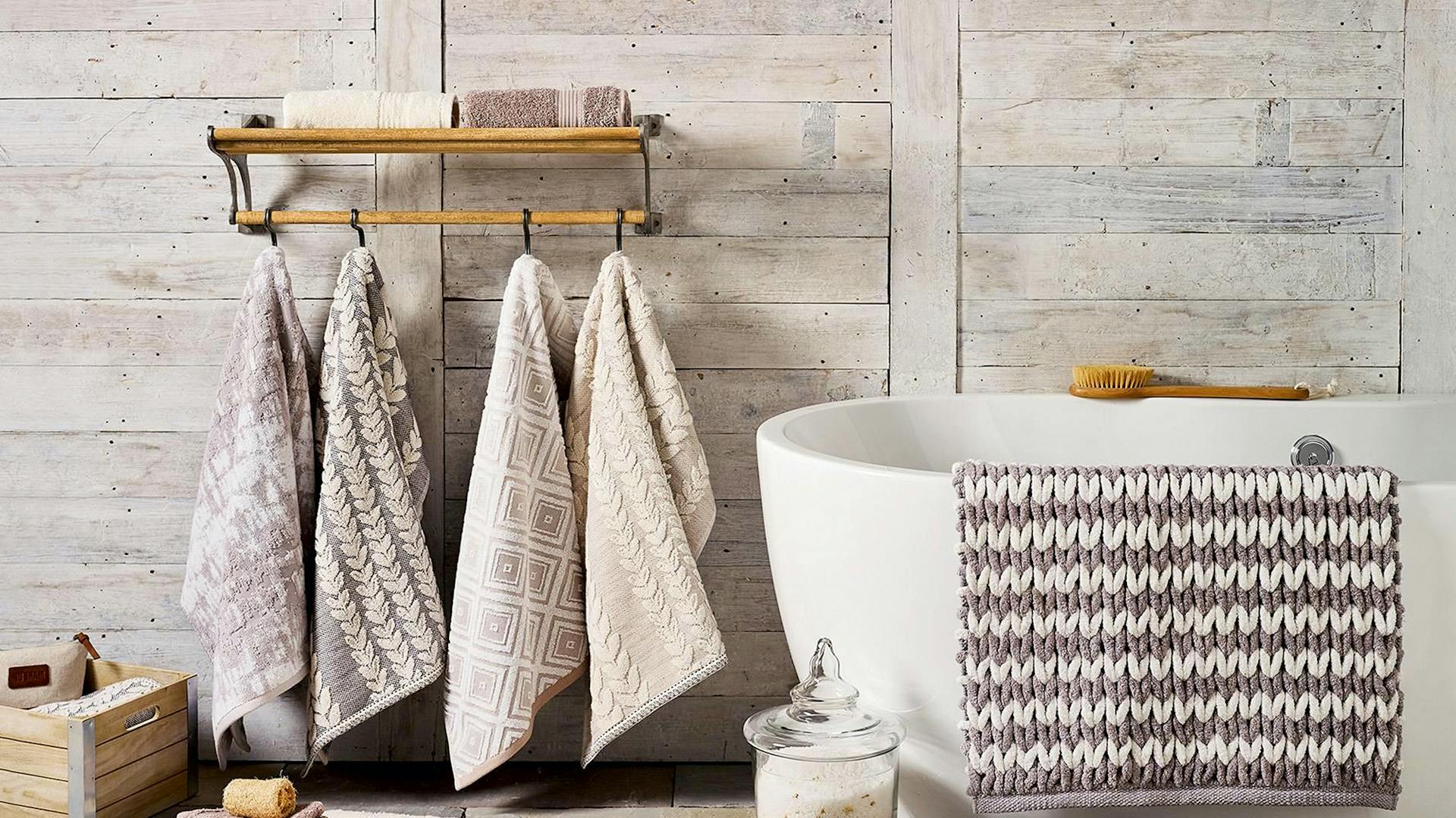 Holzwand mit Handtuchhalter. Daran hängen 4 helle Handtücher. Daneben eine weiße Badewanne.