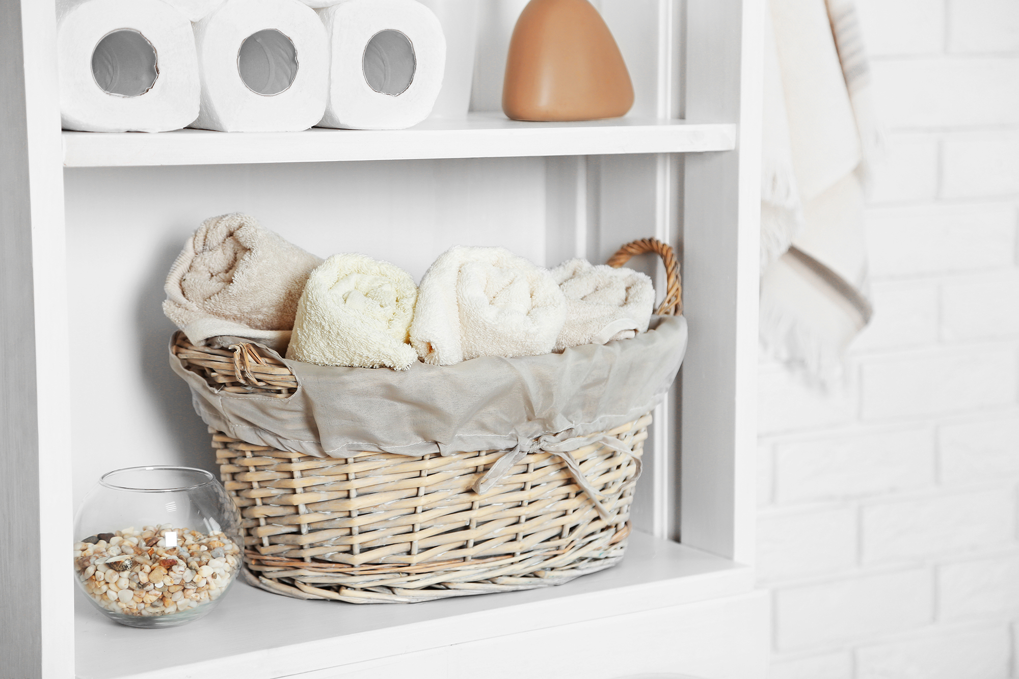 Biela polica s prúteným košíkom. Vo vnútri sú svetlé zrolované uteráky. 