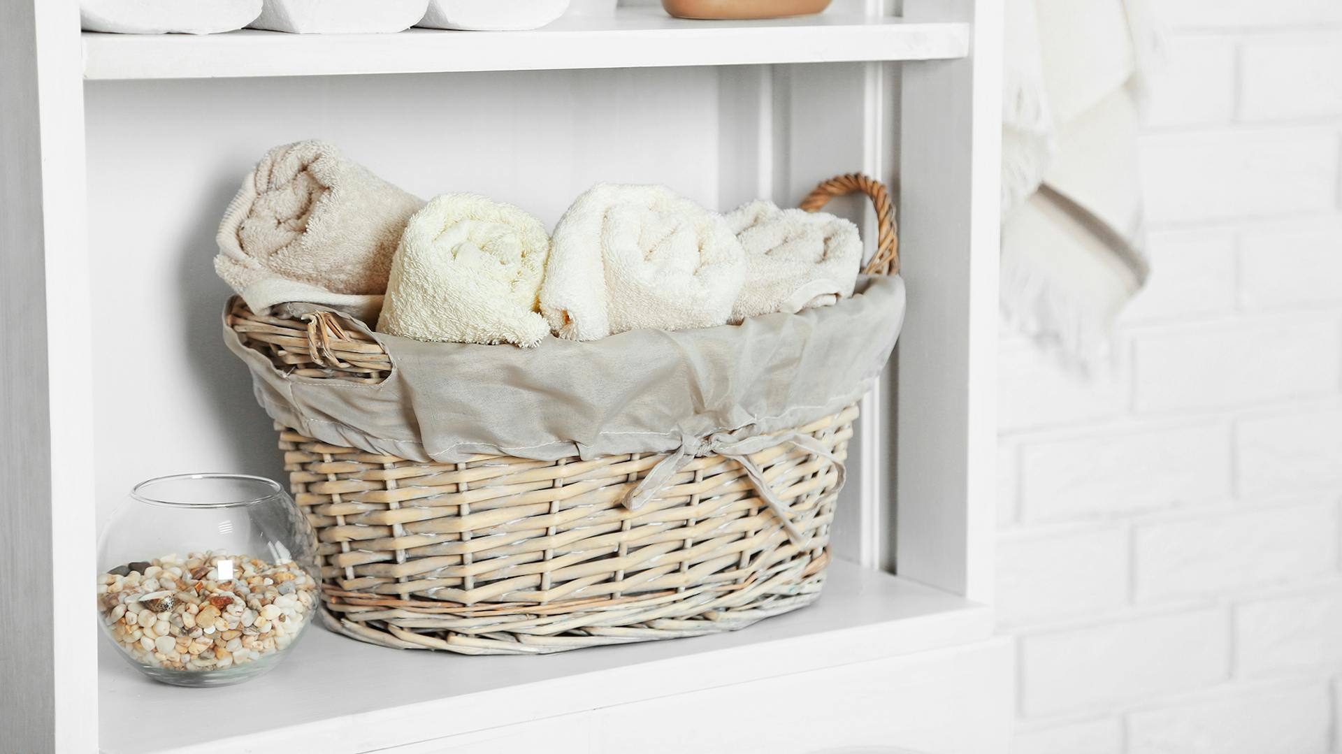 Weißes Regal mit Weidenkorb. darin liegen helle aufgerollte Handtücher.