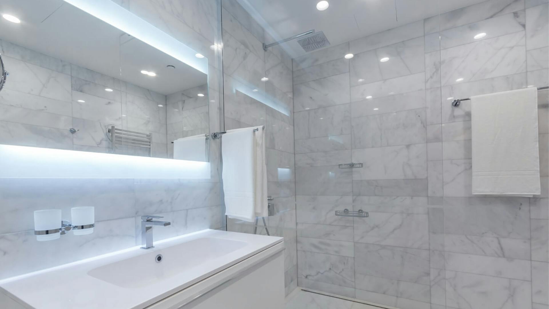 Bad mit hellgrauen Fliesen und indirekter Beleuchtung am Spiegel und in der Dusche.