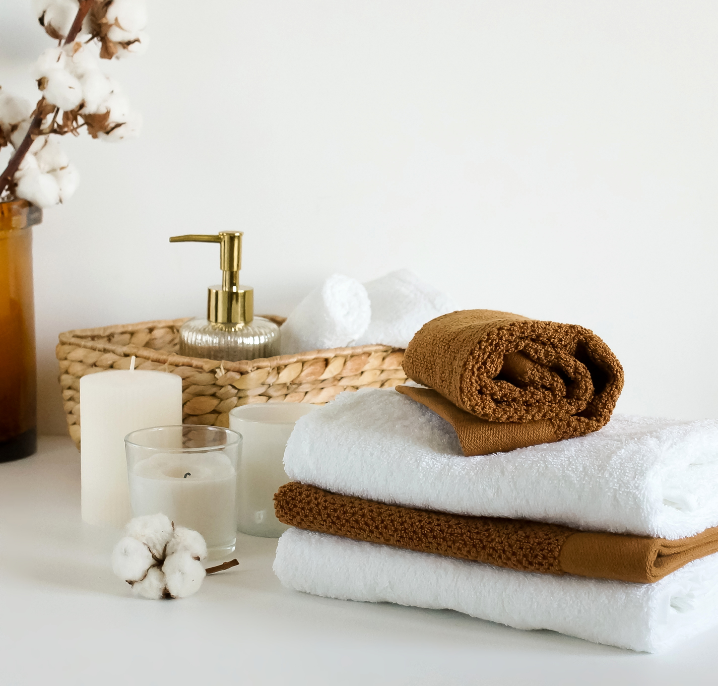 Accessori per il bagno marrone: asciugamani impilati marrone e bianco, dosatore di sapone in un cesto intrecciato. Davanti ci sono delle candele bianche.