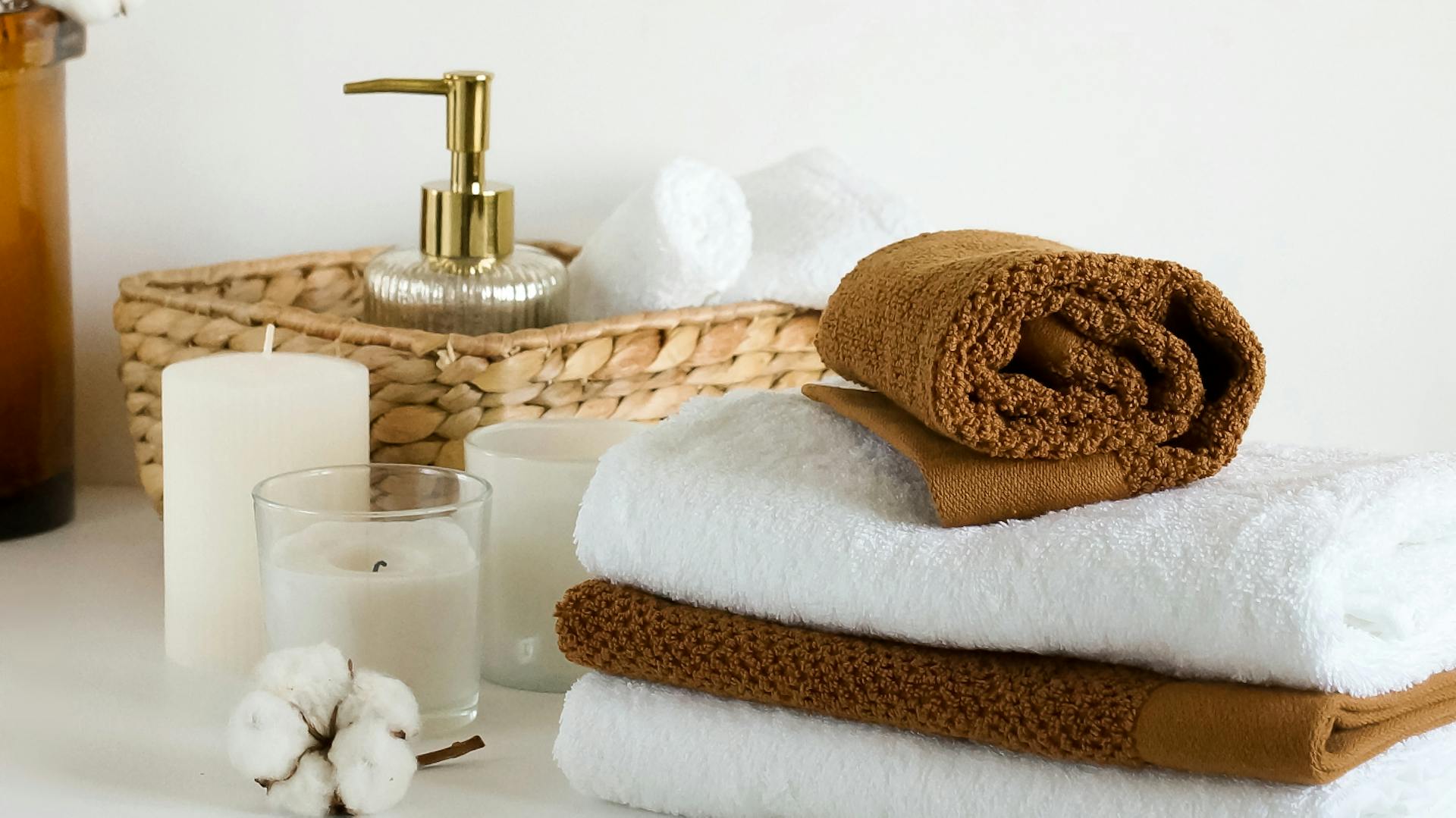 Accessori per il bagno marrone: asciugamani impilati marrone e bianco, dosatore di sapone in un cesto intrecciato. Davanti ci sono delle candele bianche.