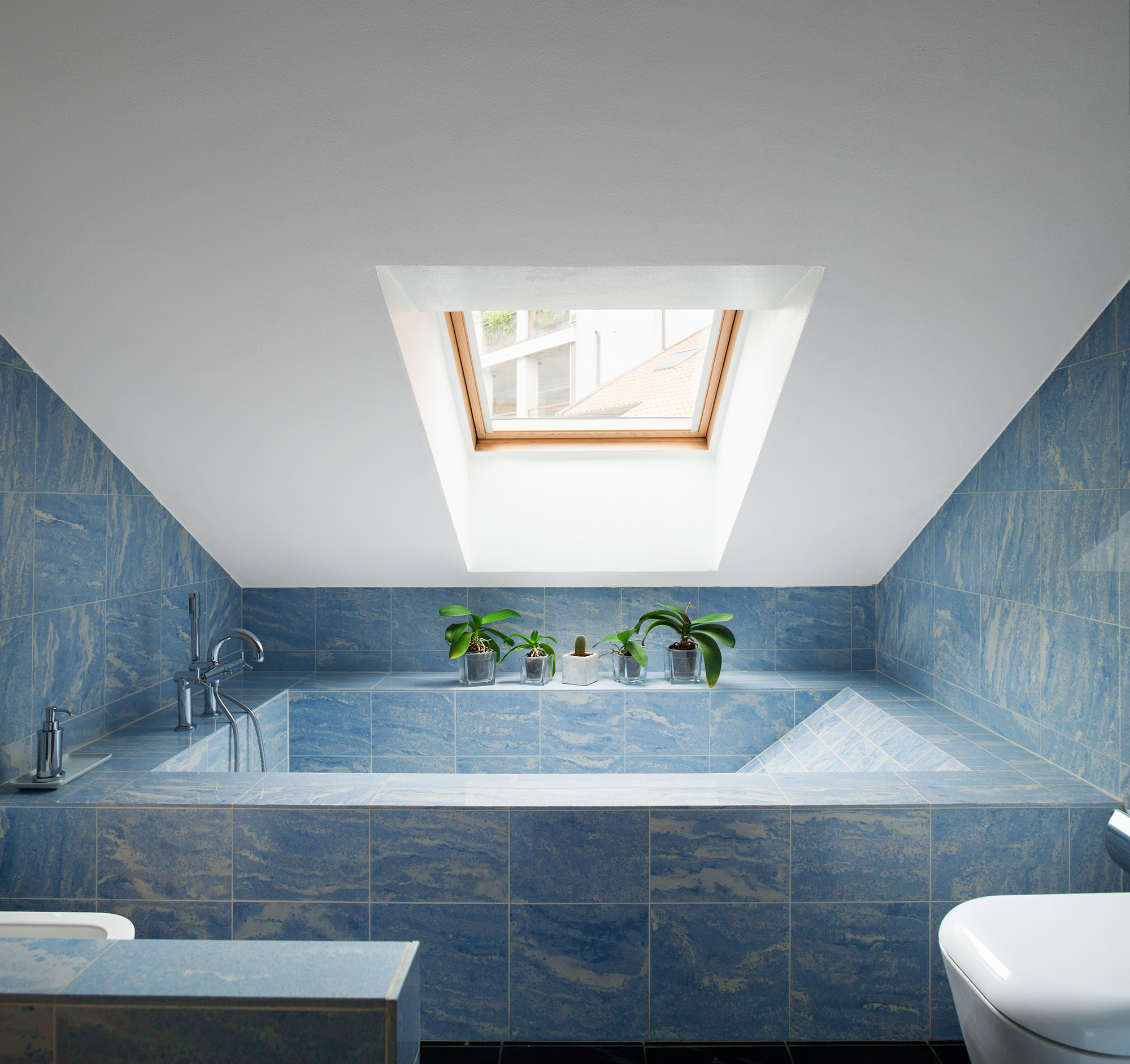 Vasca da bagno piastrellata in blu sotto un tetto spiovente verniciato in bianco con finestre.