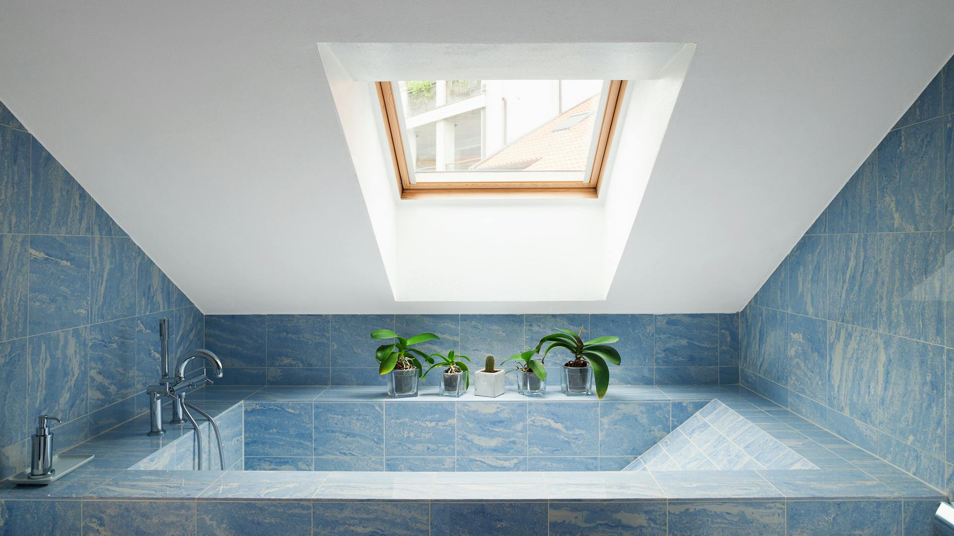 Vasca da bagno piastrellata in blu sotto un tetto spiovente verniciato in bianco con finestre.