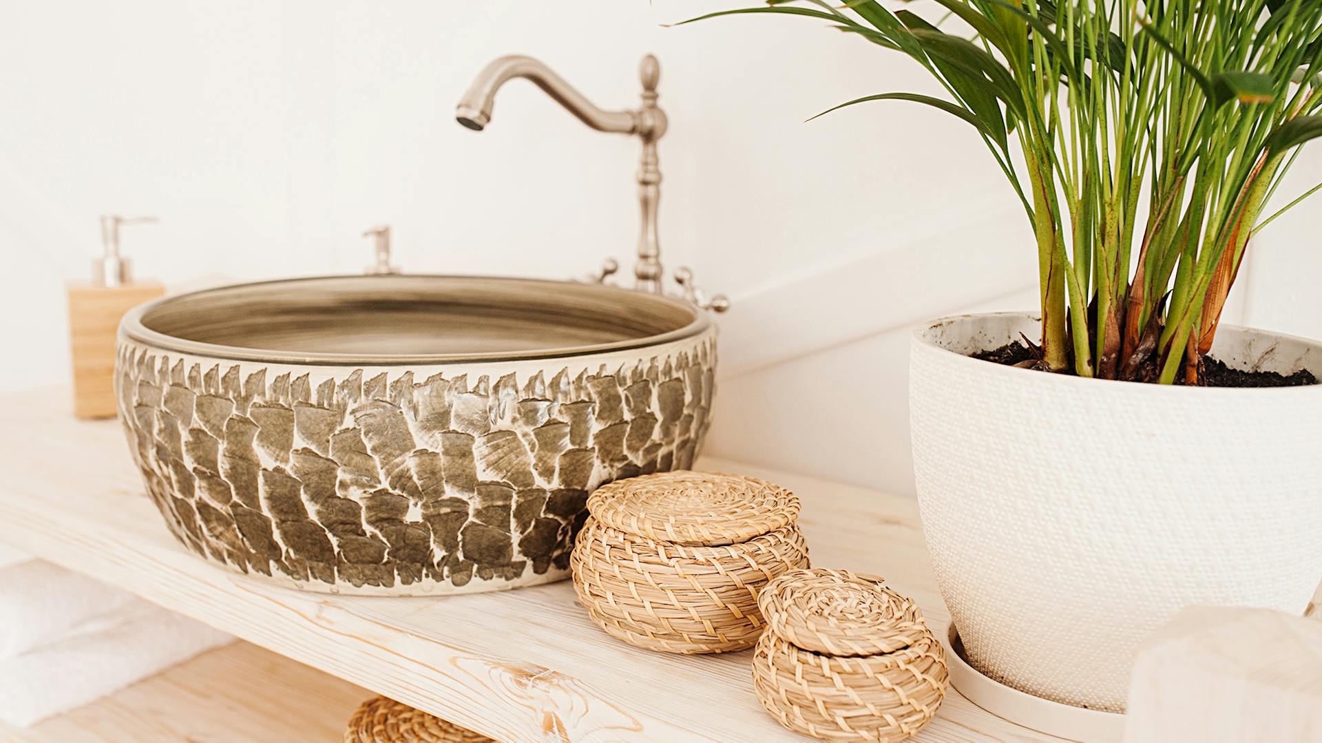 Dřevěné umyvadlo, košíky, dávkovač mýdla a pokojová rostlina na dřevěné desce pod umyvadlo.