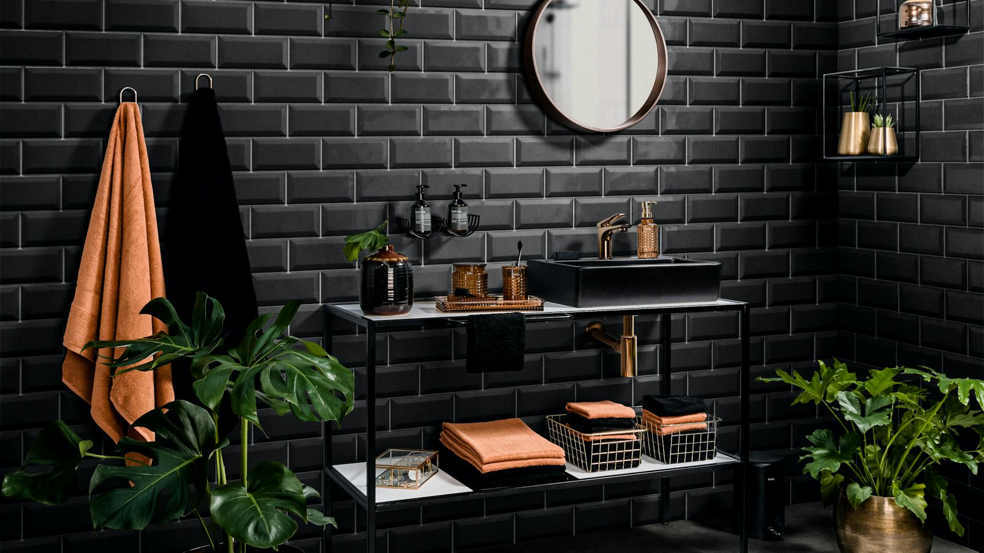 Lavabo bianco e nero con specchio davanti ad una parete in mattoni neri con piante d’appartamento, asciugamani color bronzo e neri e decorazione.