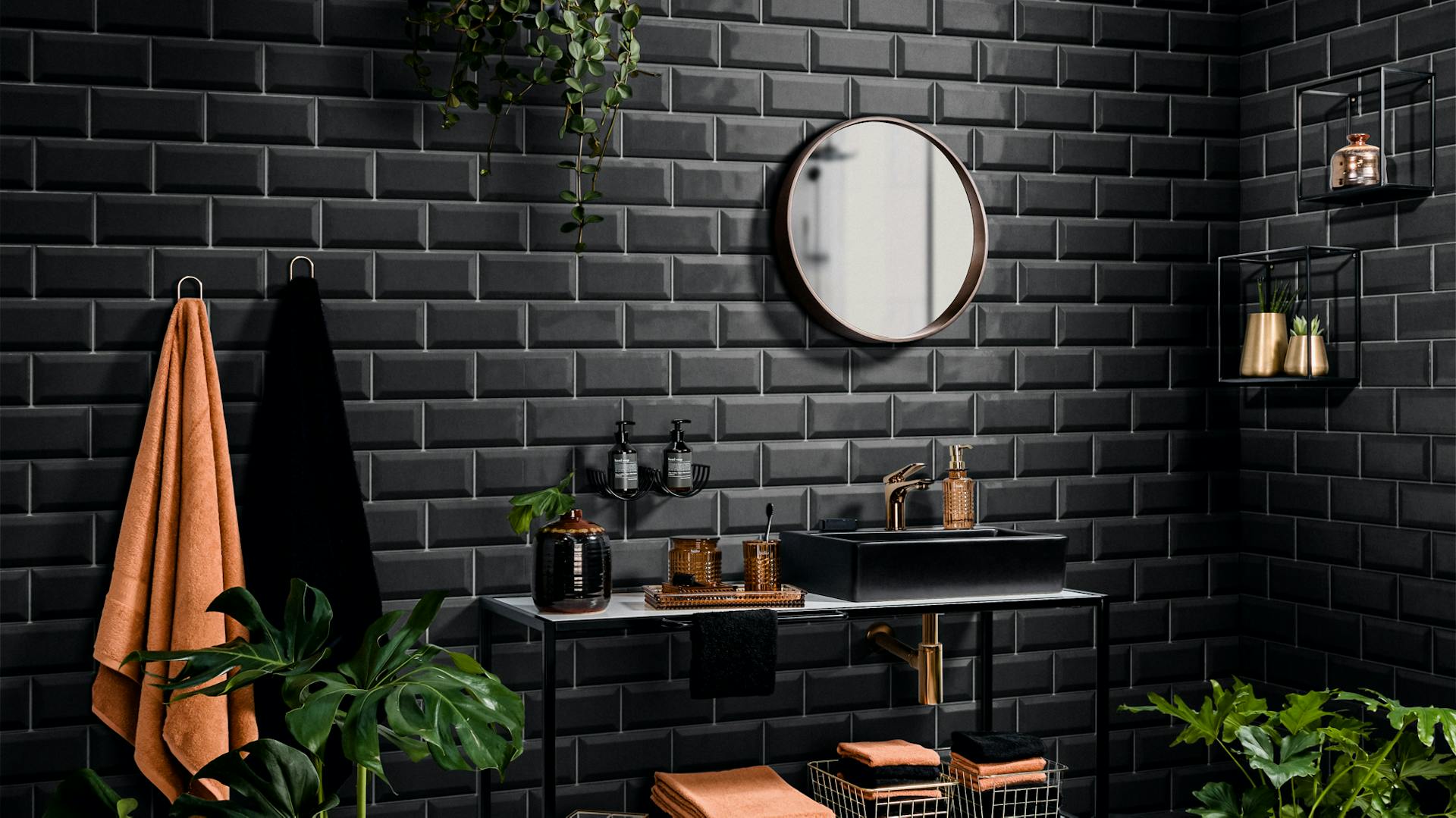 Černo-bílá deska pod umyvadlo se zrcadlem před černou cihlovou zdí s pokojovými rostlinami, ručníky v bronzové a černé barvě a dekoracemi.