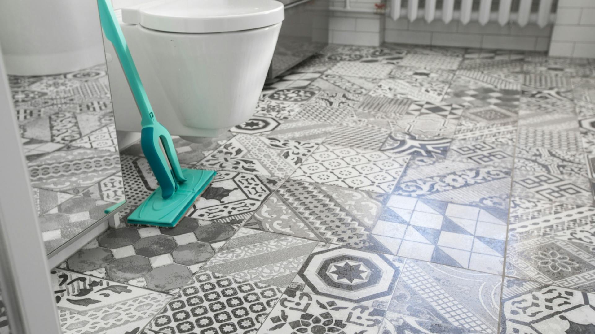 Badezimmer Boden mit Mosaikmuster auf den Fliesen. Seitlich steht ein Bodenwischer neben einer weißen Toilette.