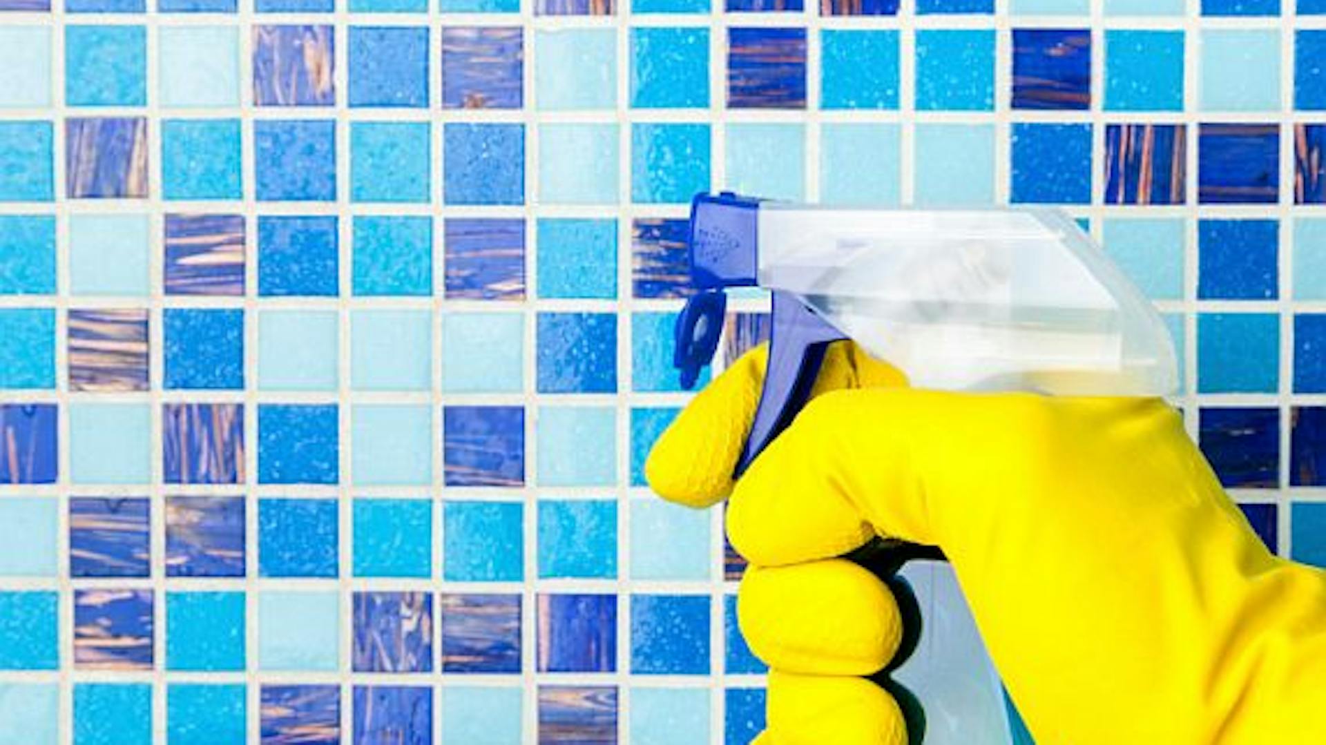 Hand mit gelbem Putzhandschuh richtet eine Sprühflasche mit Putzmittel auf blaue Mosaikfliesen.