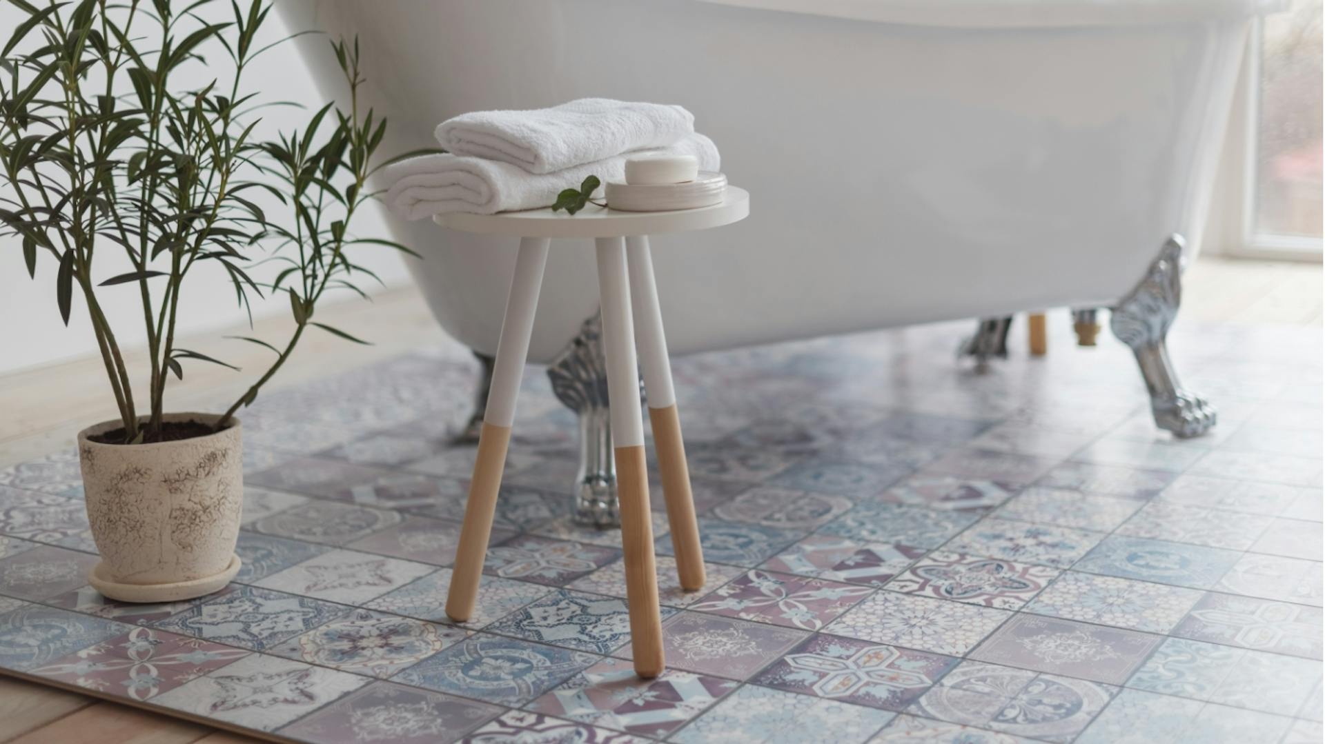 Weiße Badewanne, davor ein Hocker aus Holz. Darauf liegen kleine weiße Handtücher. Neben dem Hocker steht eine kleine grüne Pflanze.