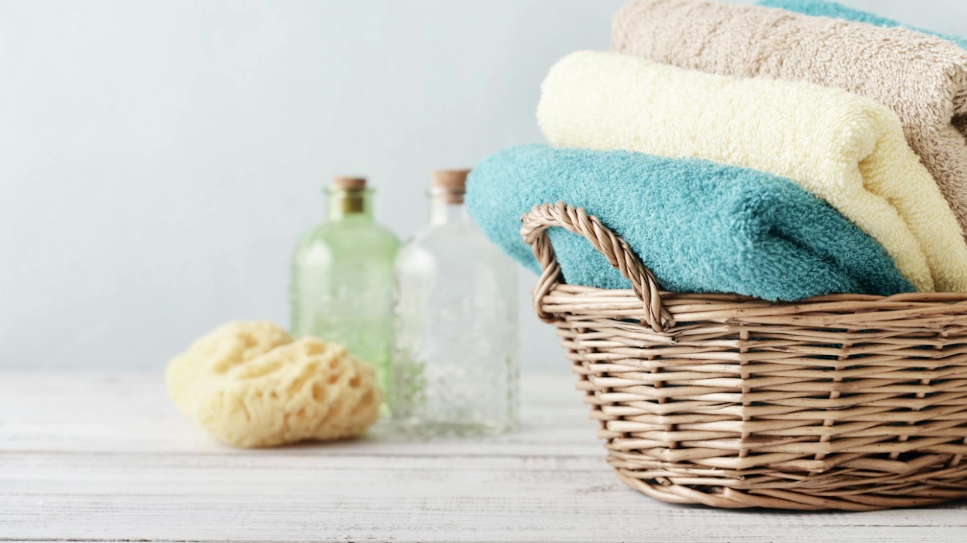 Weidenkorb mit gefalteten Handtüchern in türkis und beige. Daneben ein Schwamm und zwei Fläschchen.