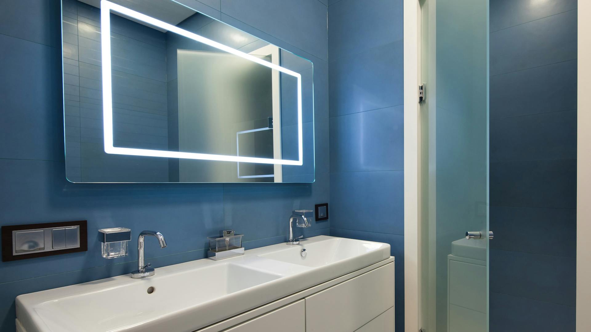 Specchio con illuminazione in un bagno con lavello chiaro e parete blu.