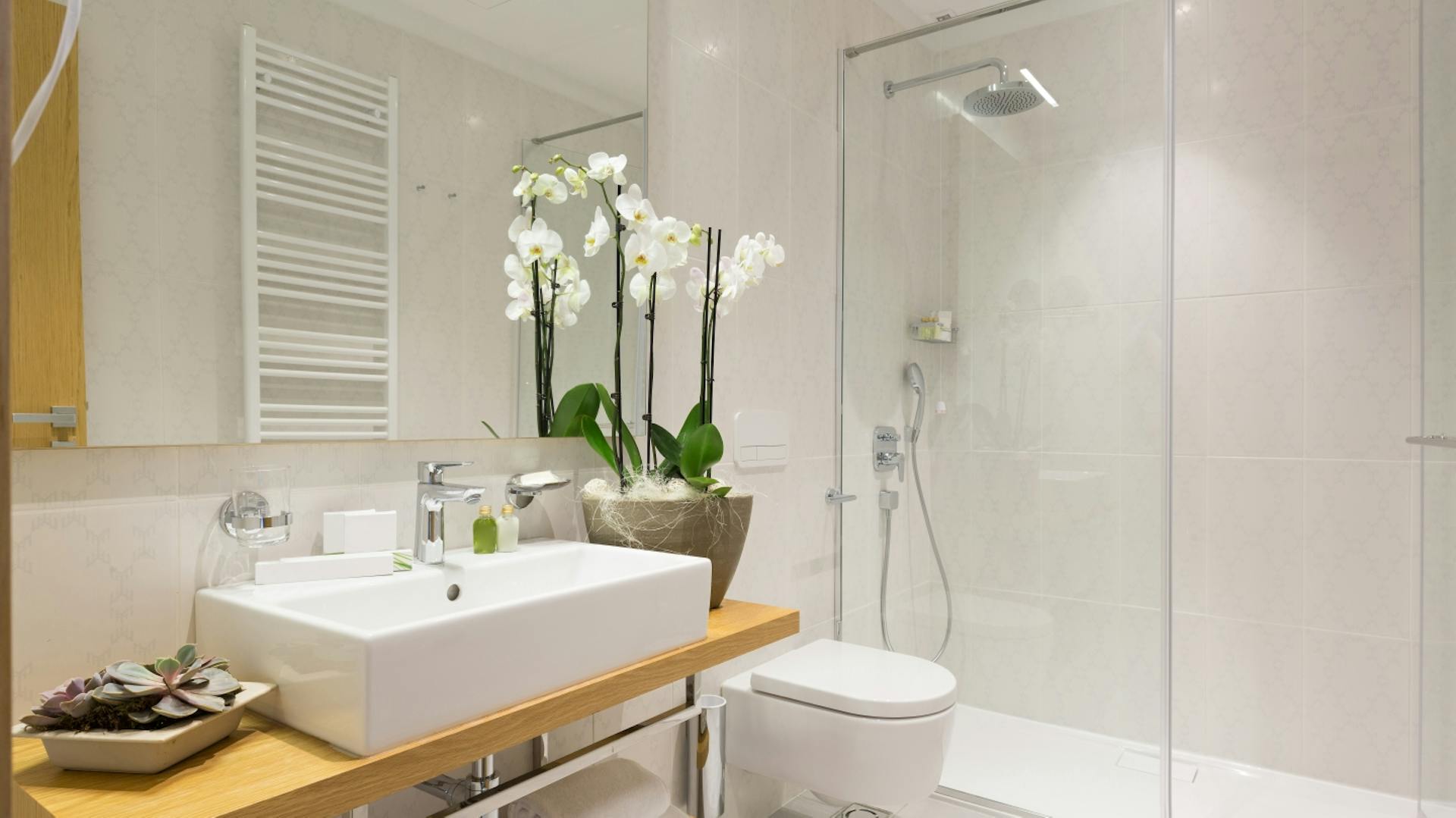 Mobile lavabo in legno ed elementi metallici, con sopra un lavabo angolare bianco. Accanto, un sanitario e una doccia walk-in. Le piastrelle chiare arrivano fino al soffitto.