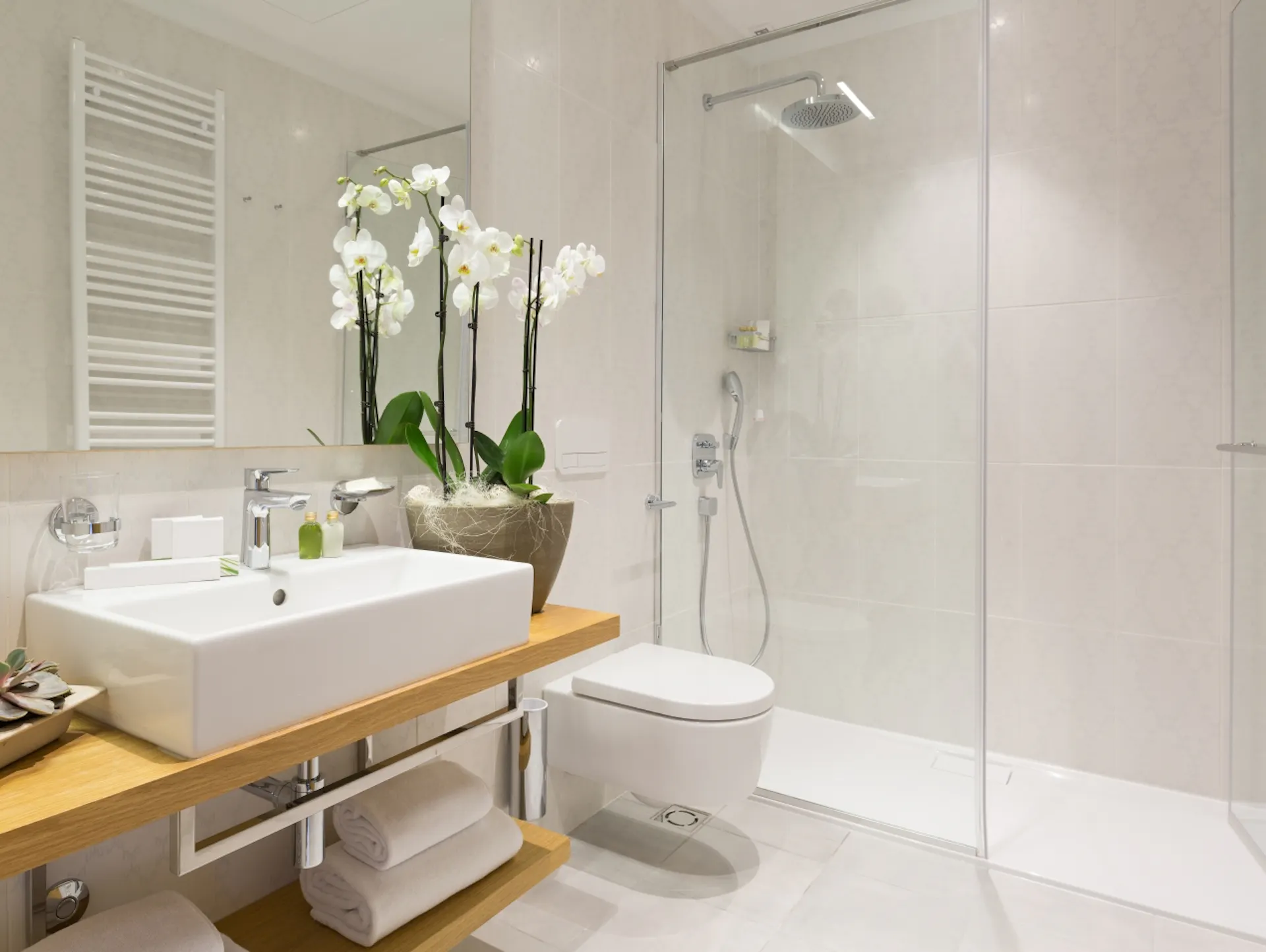 Mobile lavabo in legno ed elementi metallici, con sopra un lavabo angolare bianco. Accanto, un sanitario e una doccia walk-in. Le piastrelle chiare arrivano fino al soffitto.