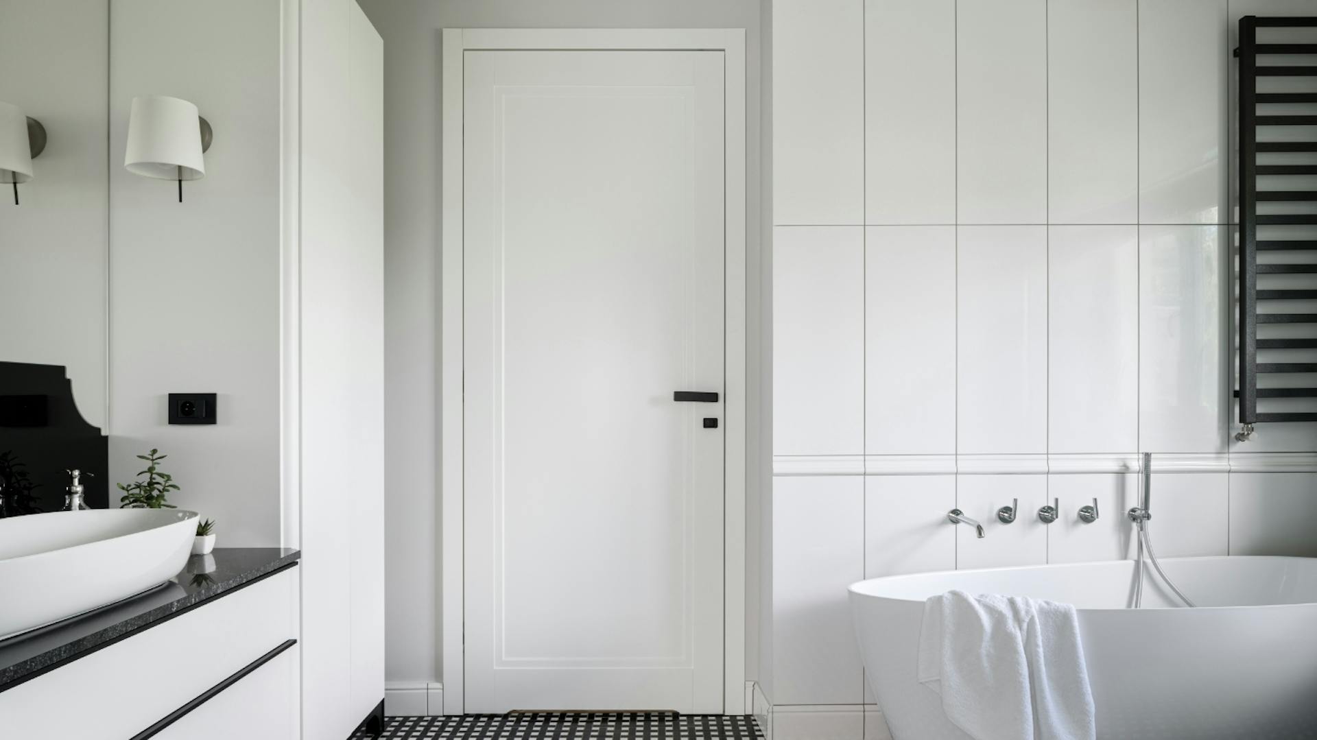 Bagno con piastrelle verticali bianche. Una vasca da bagno bianca e un mobile lavabo bianco e nero. Il pavimento è piastrellato di nero.