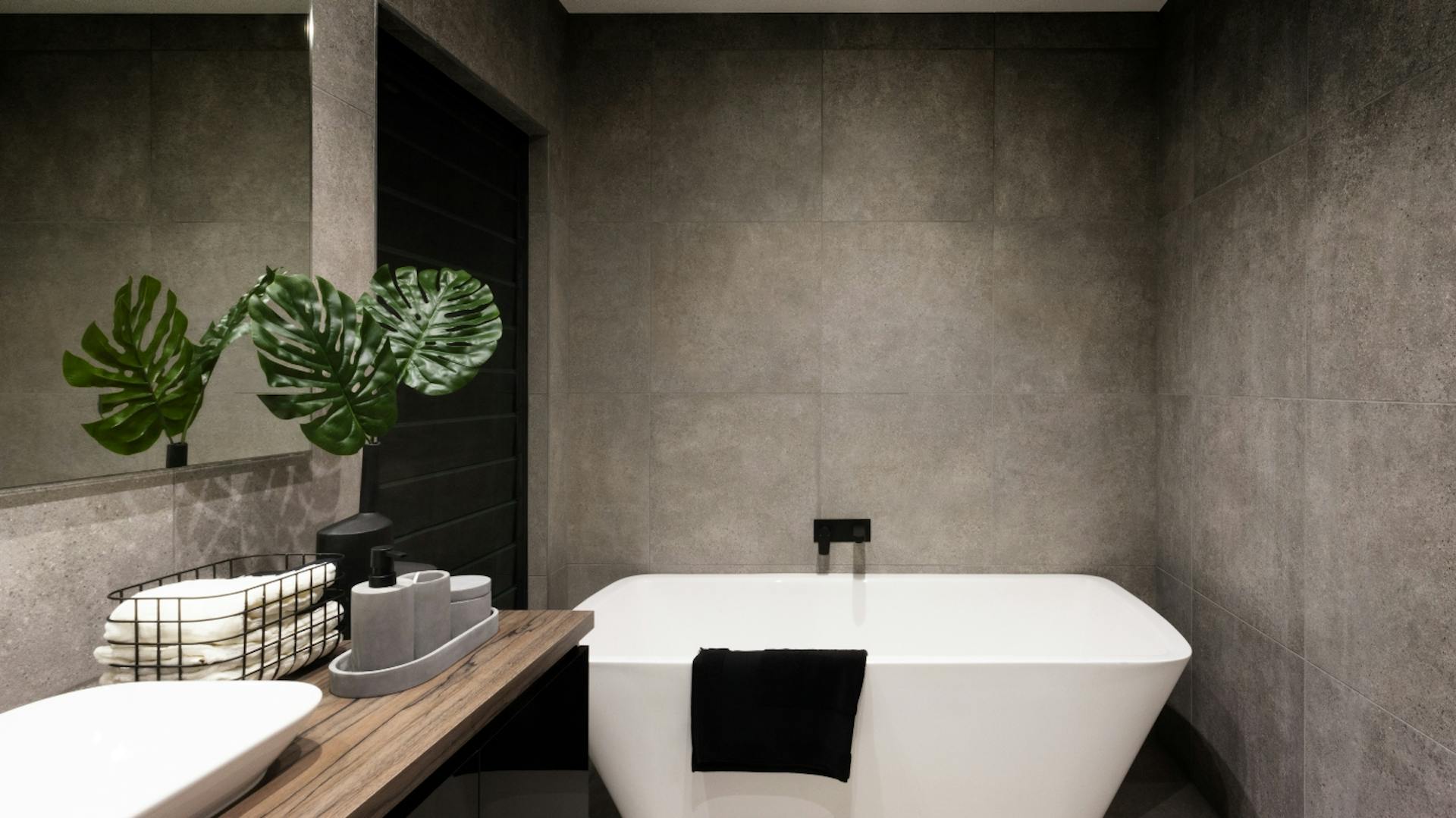 Piastrelle scure e grandi. Di fronte c'è una vasca da bagno bianca e, all'esterno a sinistra, un mobile lavabo in legno con un lavabo bianco.