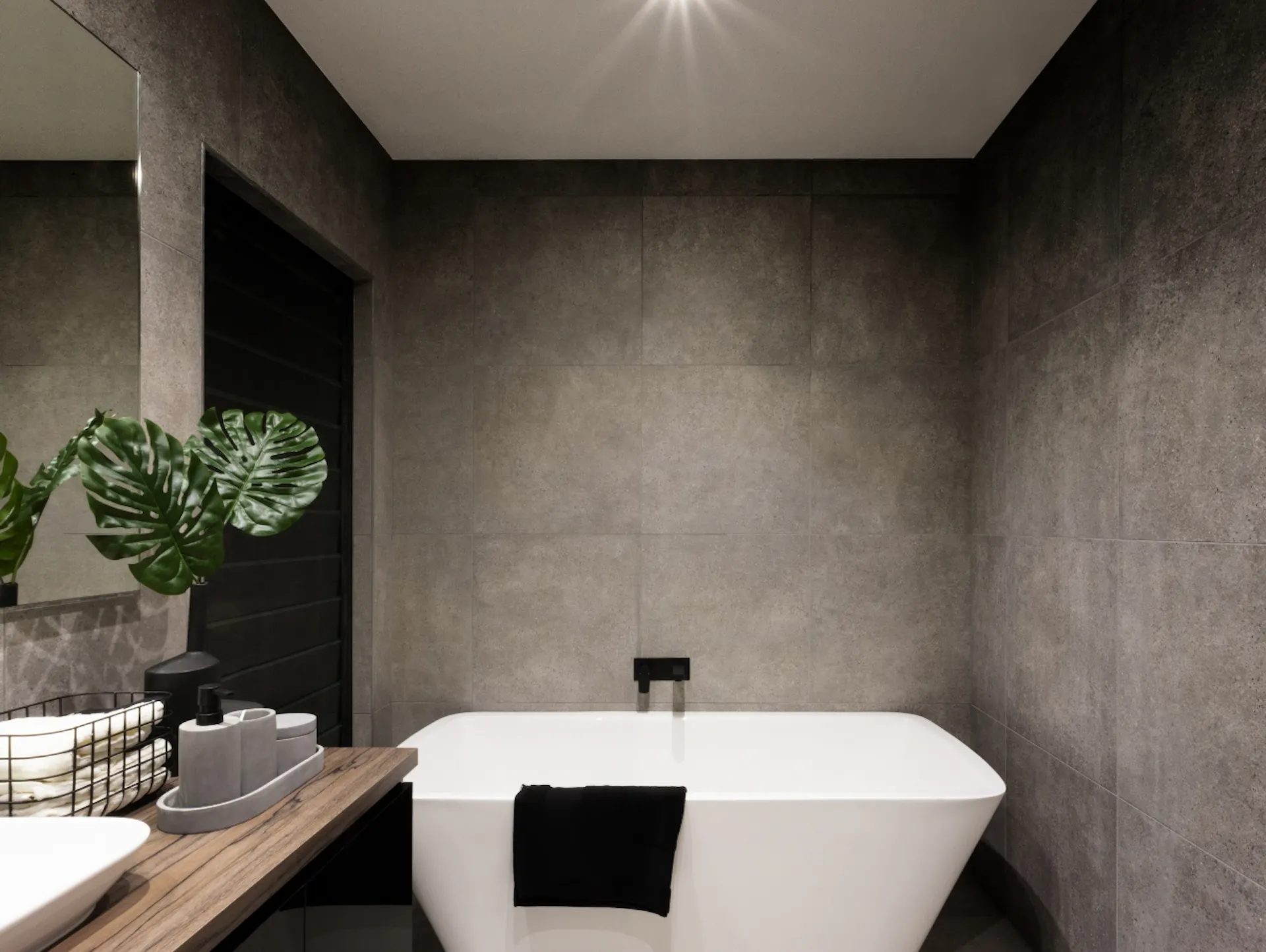 Piastrelle scure e grandi. Di fronte c'è una vasca da bagno bianca e, all'esterno a sinistra, un mobile lavabo in legno con un lavabo bianco.