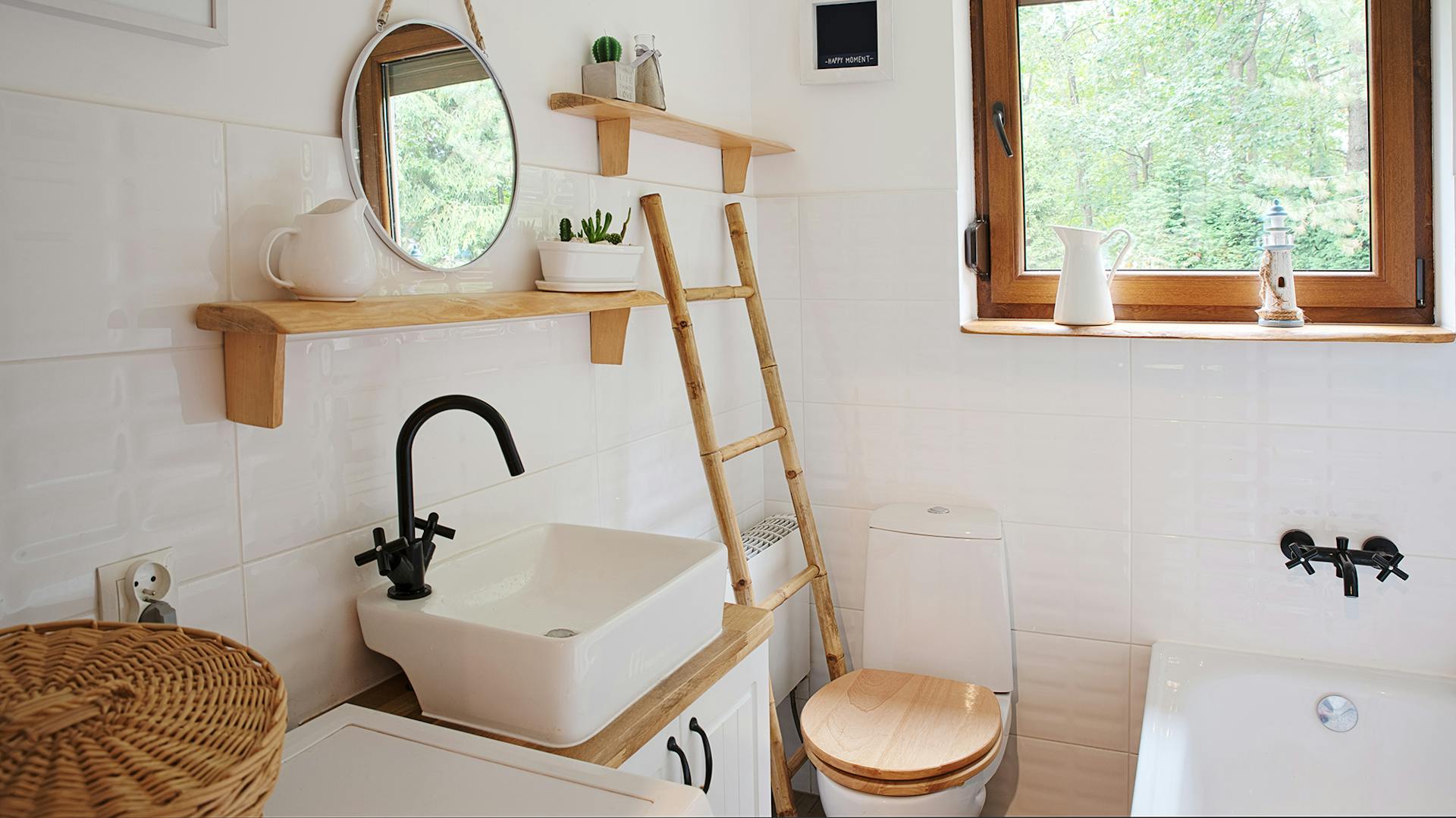 Piccolo bagno con piastrelle chiare, lavabo bianco con rubinetto nero. Il sedile WC, gli elementi decorativi e i telai delle finestre sono in legno.