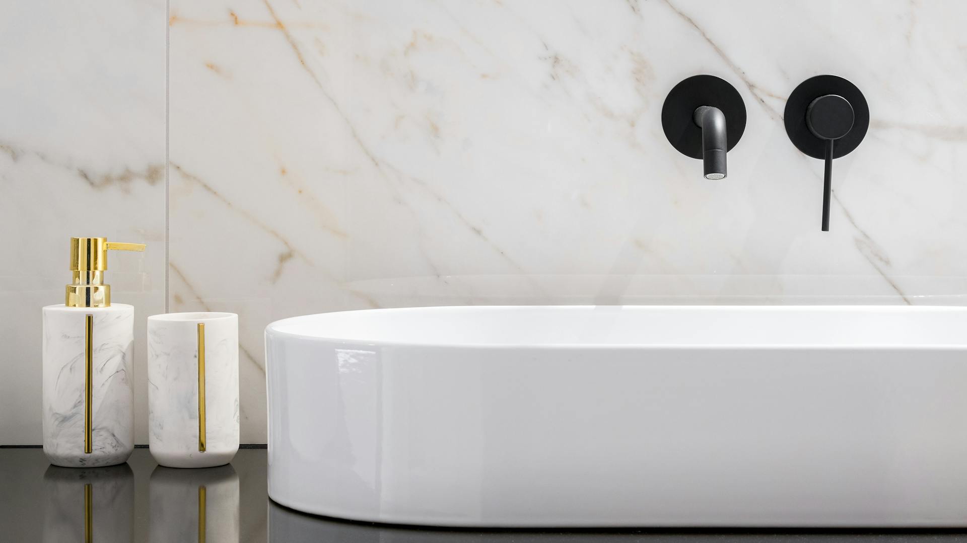 Schwarze Oberfläche eines Waschtischs, darauf weißes modernes Waschbecken mit schwarzen Armaturen an einer Mamorwand.