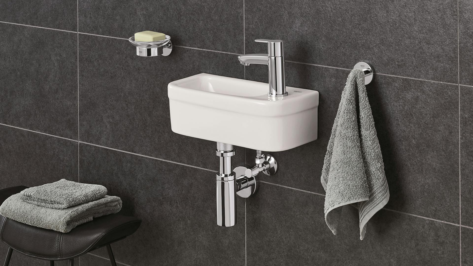 schmales Waschbecken für Schlauchbad an dunkel gefliester Wand neben Handtuch Schemel und Seifenhalter