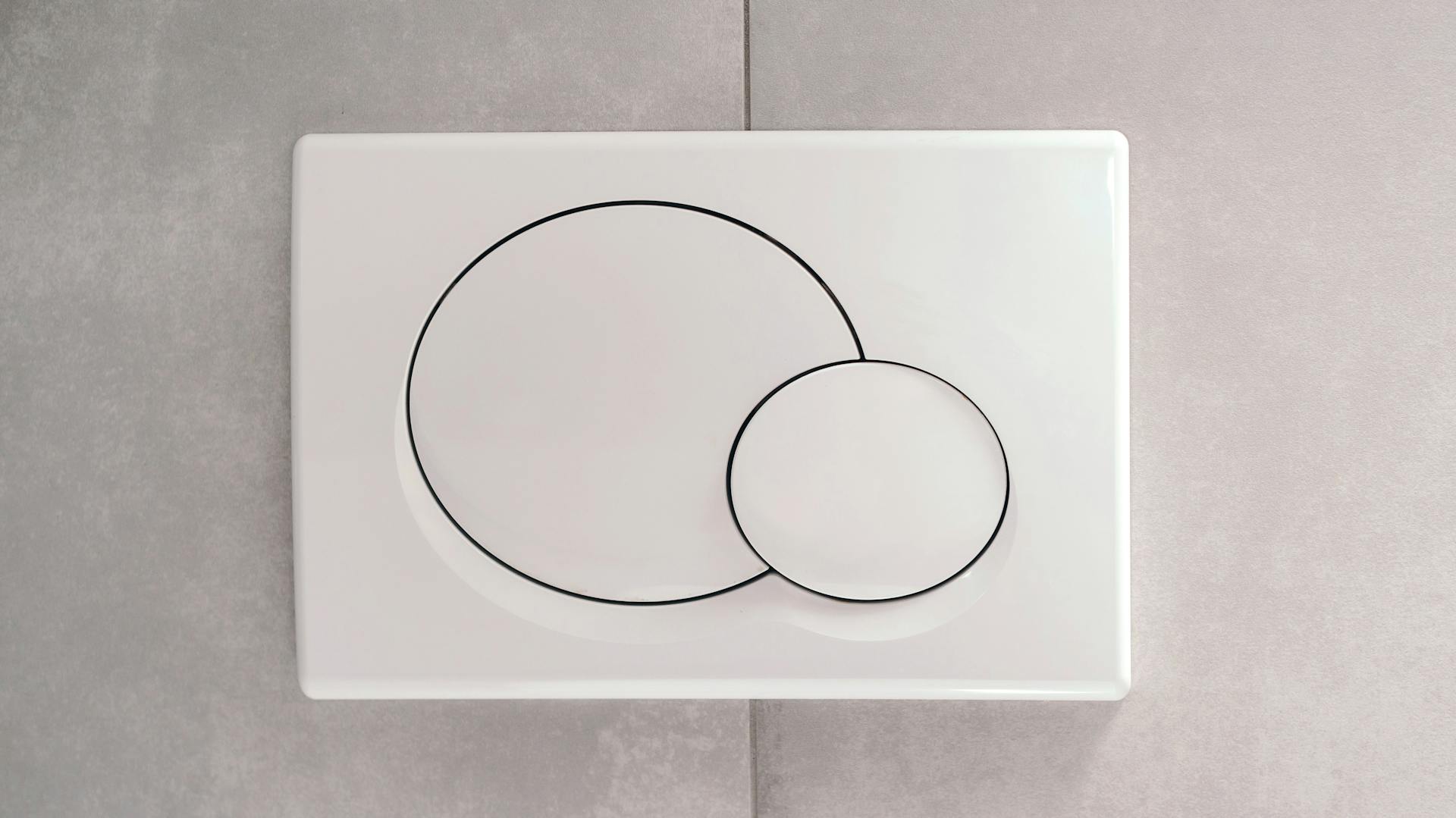 Weiße Spültaste eines Unterputz-WCs auf grauen Wandfliesen.