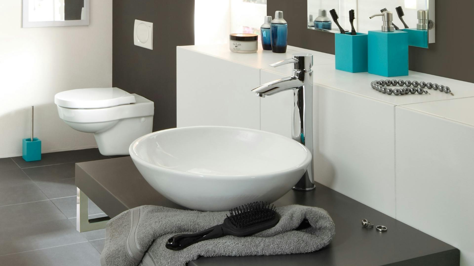 Weißes, rundes Aufsatzwaschbecken mit silberne Armatur neben grauem Handtuch und schwarzer Bürste auf dunklem Waschtisch.