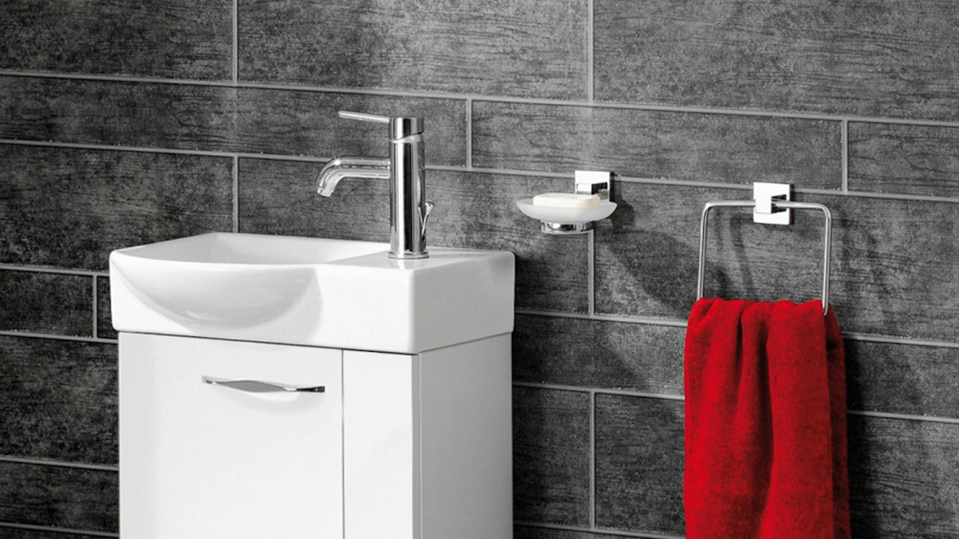 Kleines, eckiges, weißes Waschbecken mit silberne Armatur auf weißem Unterschrank neben rotem Gästehandtuch und Seife für Gäste-WC.