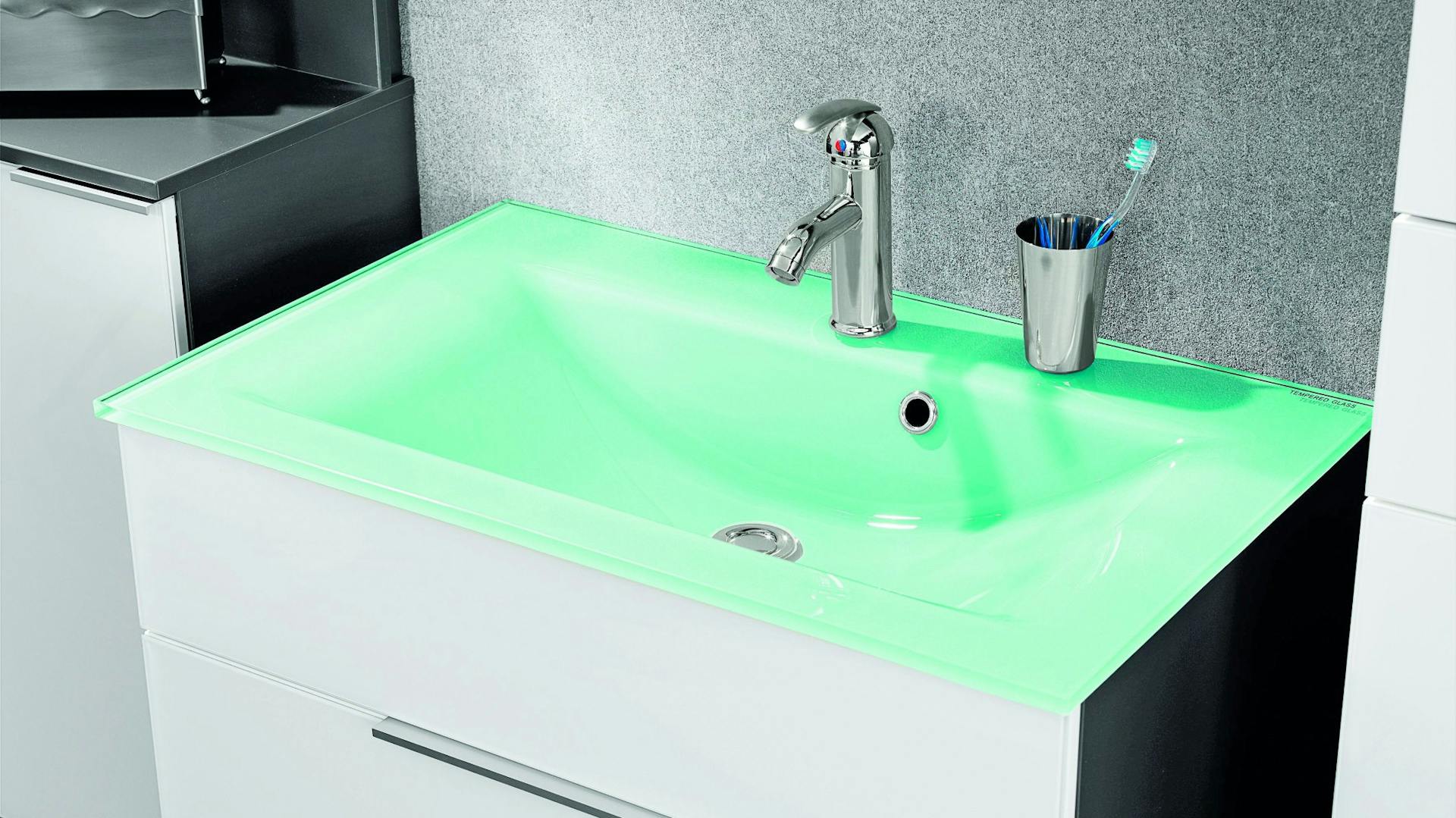 Kleines grünes Waschbecken aus Glas mit silberner Armatur und Zahnputzbecher auf Unterschrank mit weißen Schubladen.