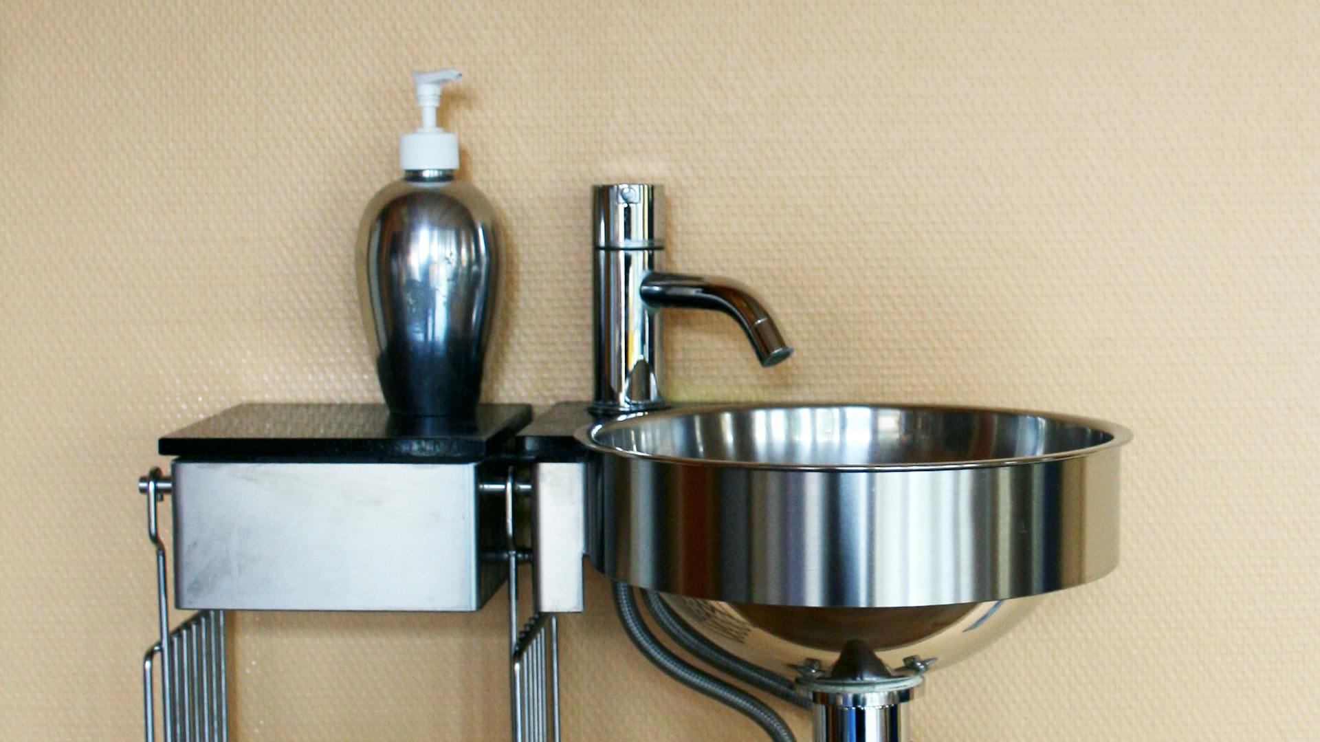 Waschbeckenset aus Stahl mit Seifenspender vor beiger Wand.