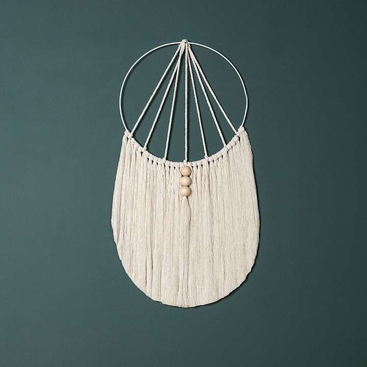 Ein weißer Wandbehang aus Makramee verziert mit kleinen Holzkugeln vor einer dunkelblauen Wand.