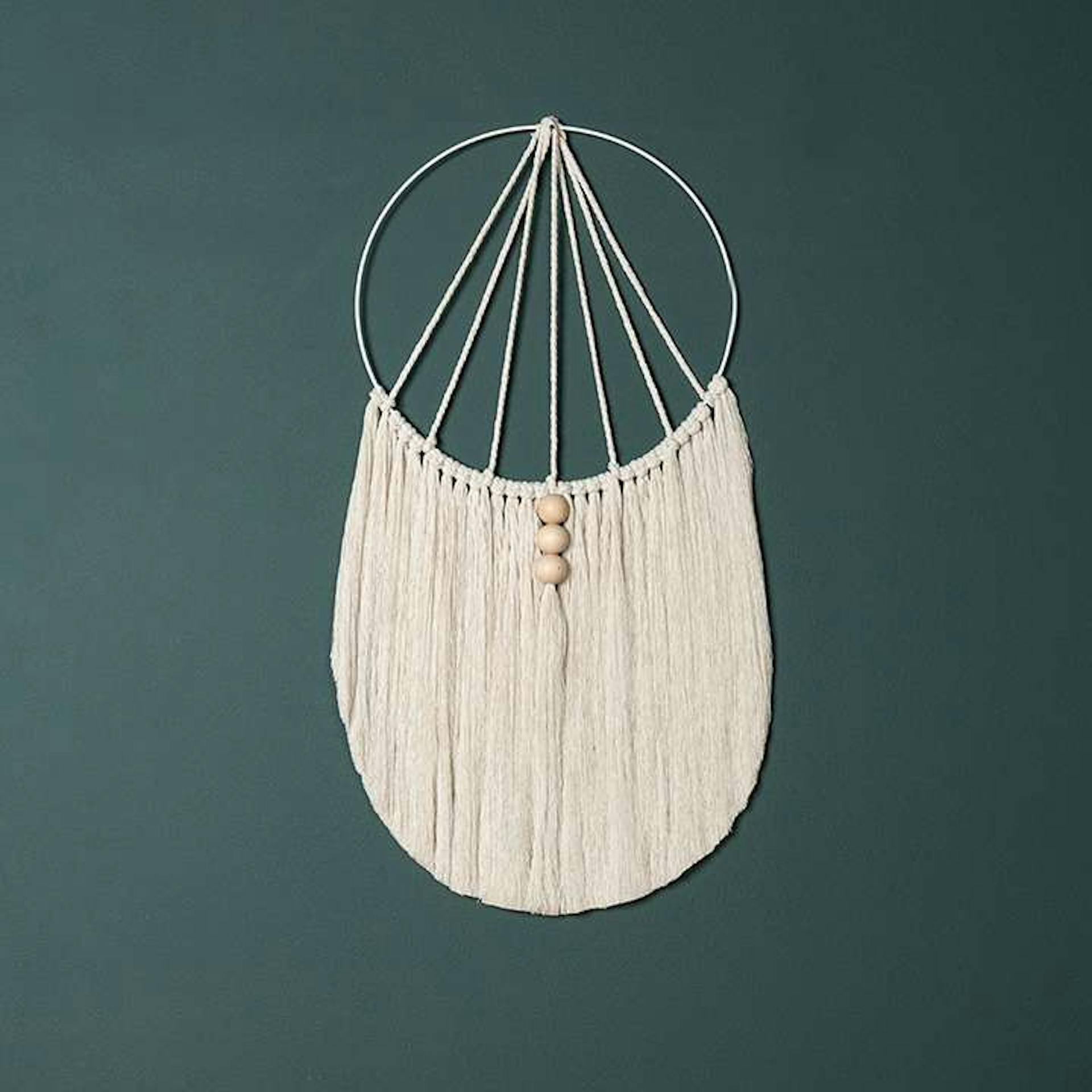 Ein weißer Wandbehang aus Makramee verziert mit kleinen Holzkugeln vor einer dunkelblauen Wand.