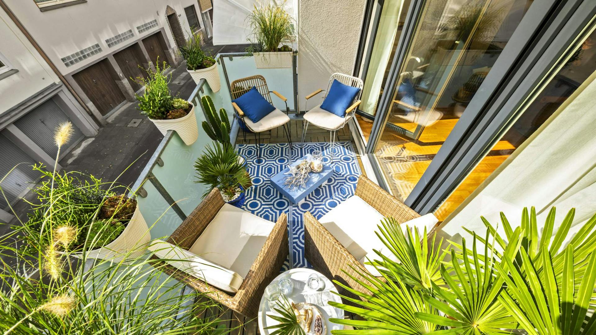 Balkon mit vielen Pflanzen und Korbstühlen mit blauen und weißen Kissen drauf.