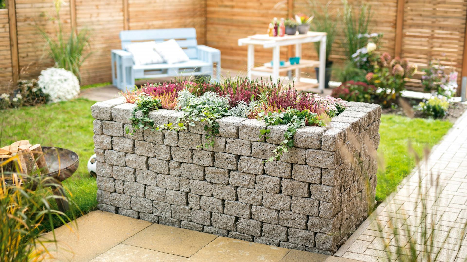 Kőből készült magaságyás homokkő teraszon, egy festői kertben világoskék paddal, fehér asztallal és fakerítéssel