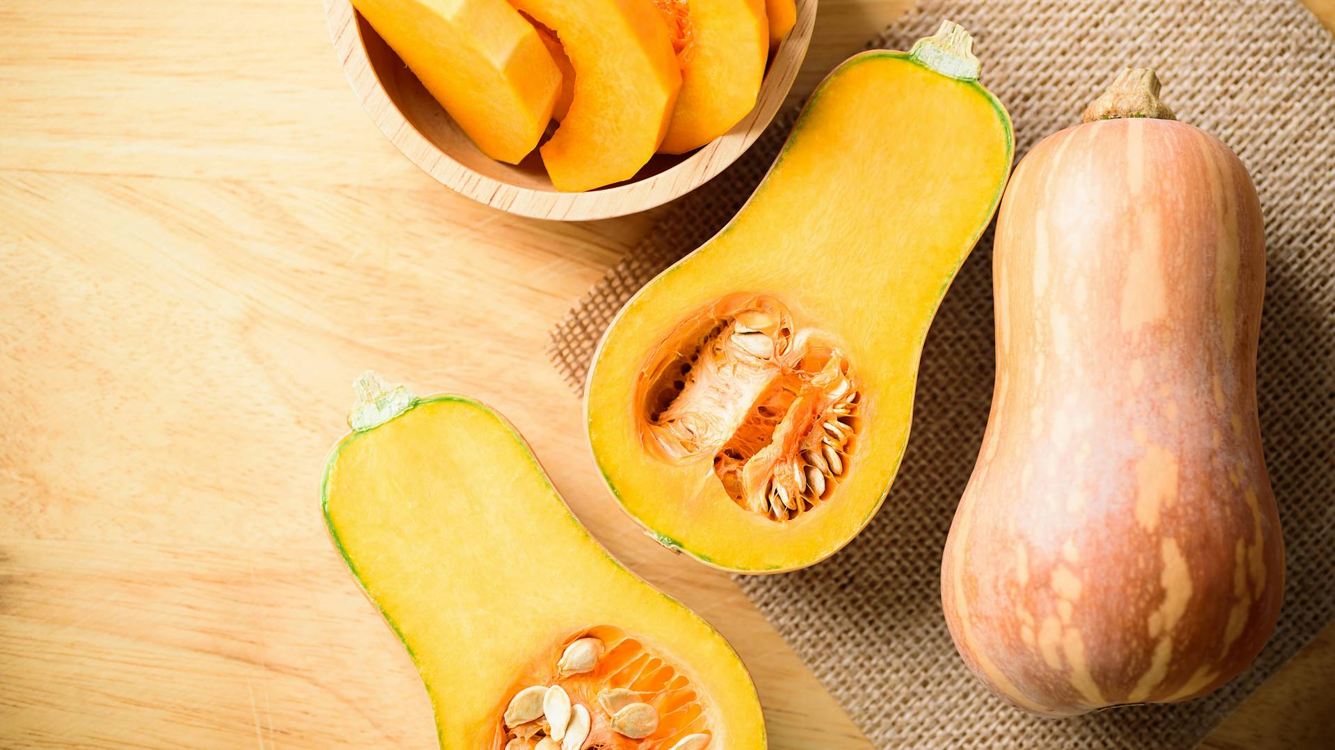 Ein aufgeschnittener Butternut-Kürbis, ein ganzer Butternut-Kürbis und Butternut-Scheiben auf einem TIsch.