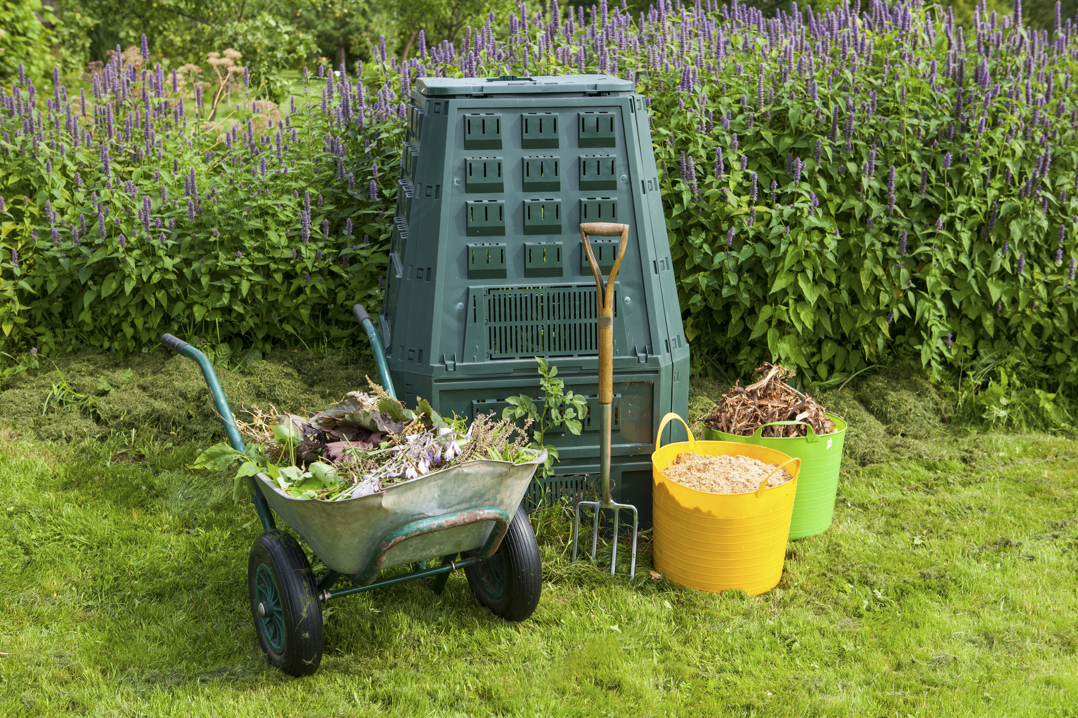 Thermokomposter im Garten, davor eine Schubkarre und zwei Eimer mit Gartenabfällen sowie eine Grabegabel.