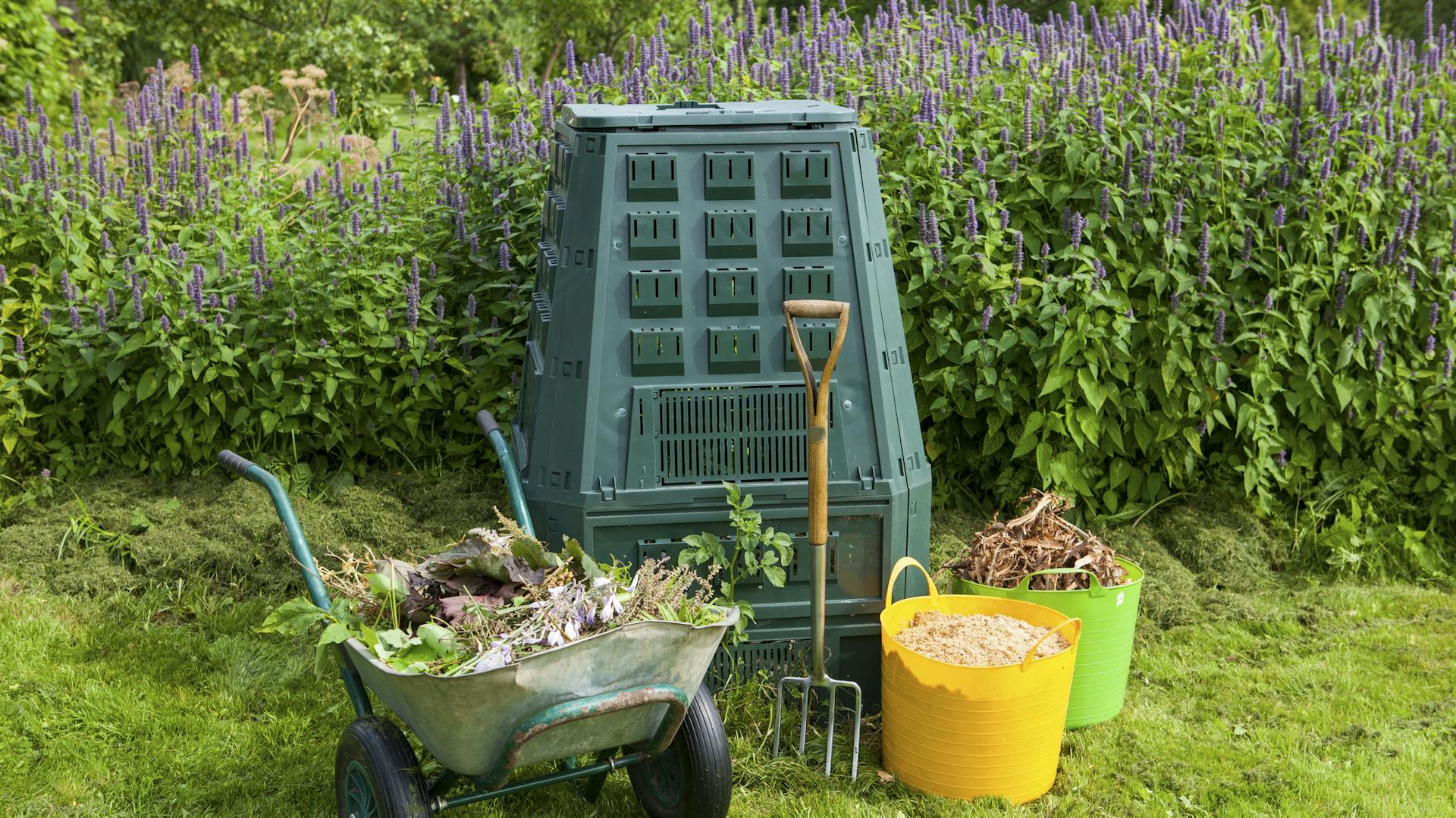Thermokomposter im Garten, davor eine Schubkarre und zwei Eimer mit Gartenabfällen sowie eine Grabegabel.