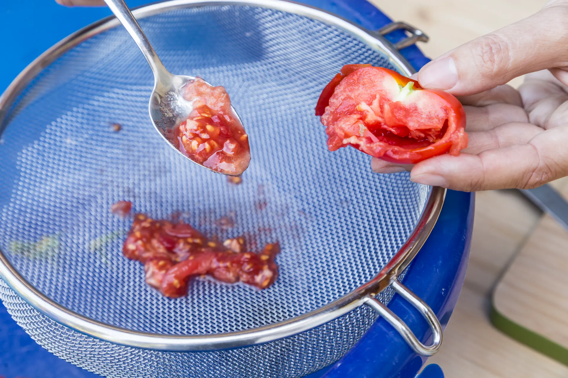 Eine Person gibt das Fruchtfleisch einer Paradeiser in ein feinmaschiges Sieb, um die Samen davon zu trennen.