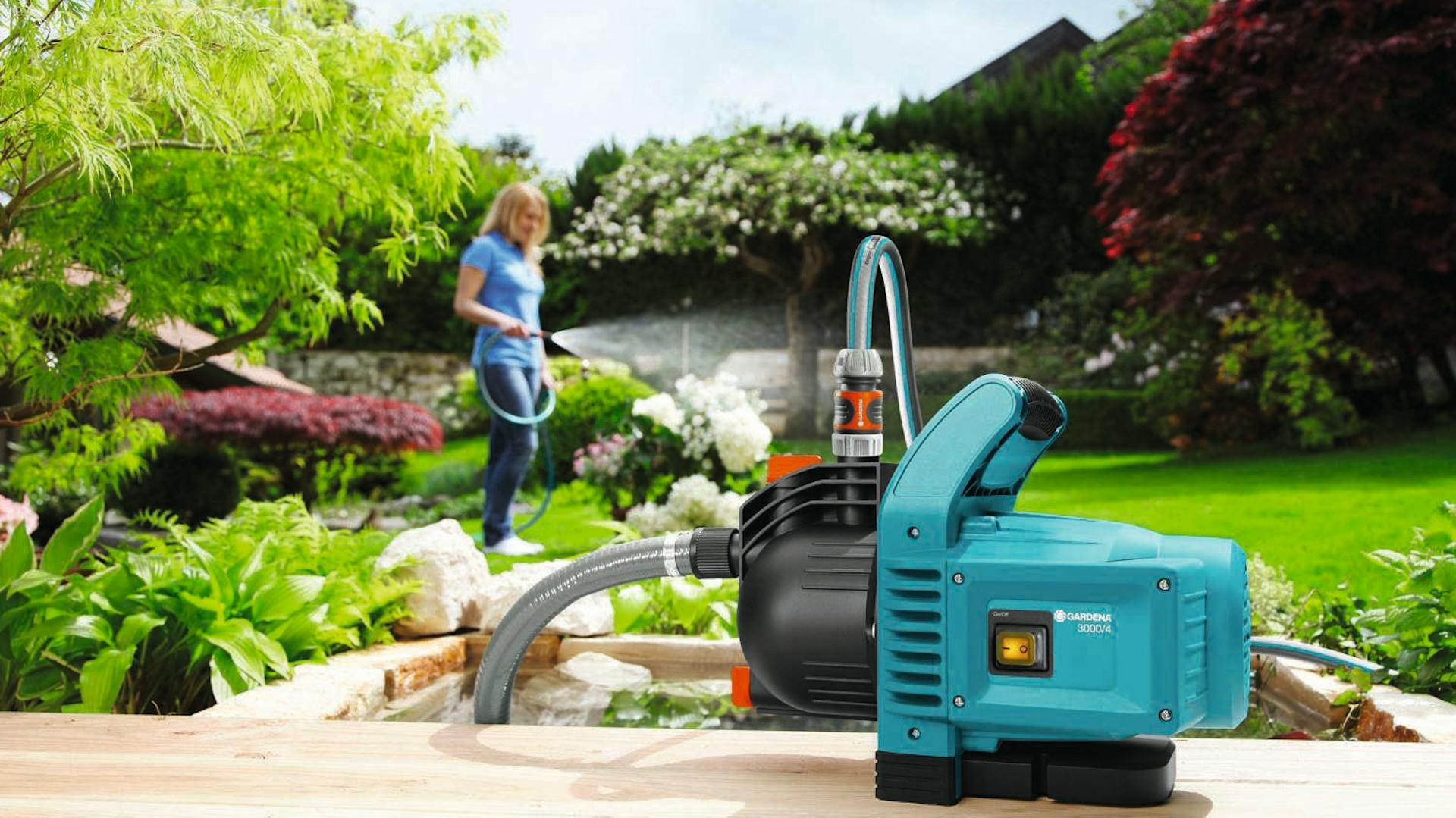 Pompa per il giardino accanto alla cisterna per l’acqua in giardino, con donna sullo sfondo che innaffia piante e prato